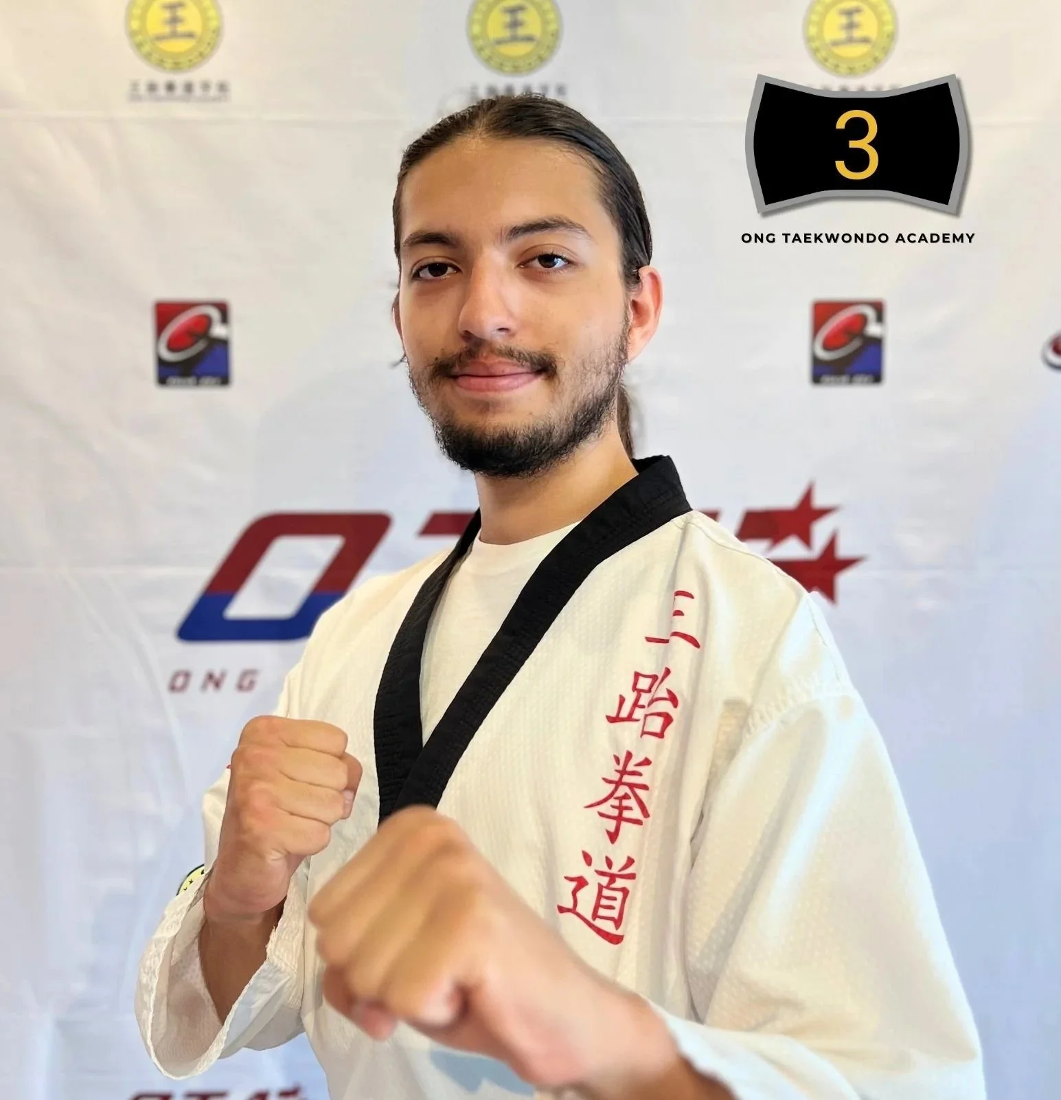 Young man in a martial arts uniform with Korean characters, standing in front of a backdrop with logos and text, posing with fists raised, showcasing his black belt.
