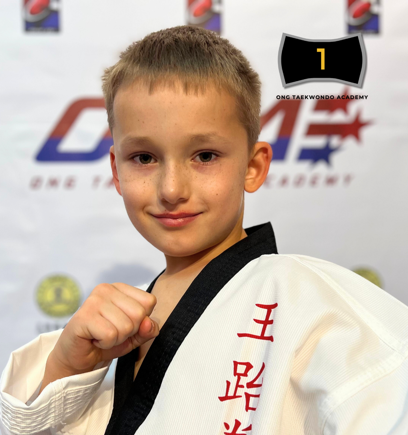 Young boy in karate uniform with black belt, posing with a fist, in front of a backdrop with sports logos, celebrating winning a black belt in Taekwondo in 2024.