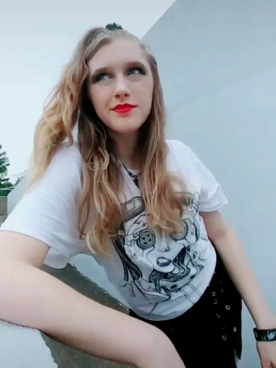 Young woman with wavy red hair, wearing a white graphic t-shirt and black shorts, posing outdoors near a white wall with a cloudy sky in the background.