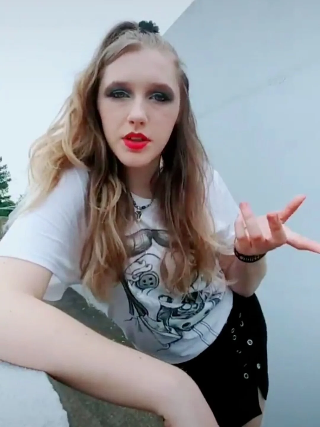 Young woman with long wavy blonde hair and makeup, wearing a graphic white t-shirt and black skirt, gesturing with her hand outdoors.