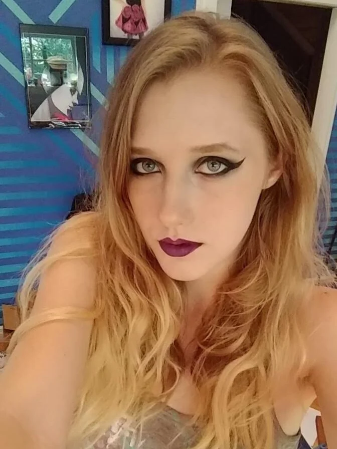 A woman with long, wavy blonde hair and bold makeup, including dark lipstick and dramatic eye makeup, is taking a selfie indoors. The background features artwork and a blue wall with diagonal stripes.