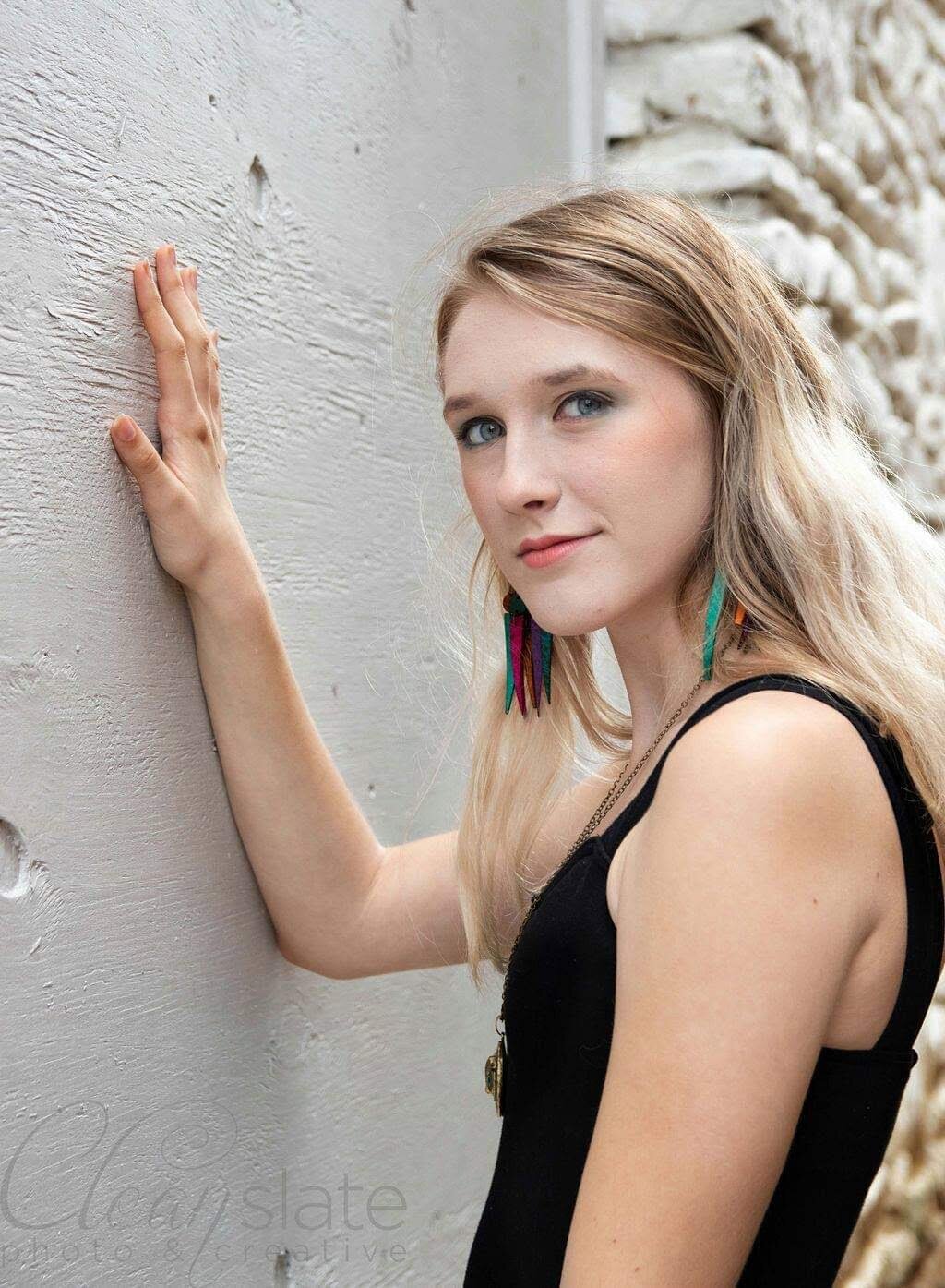 A young woman with long blonde hair, wearing colorful feather earrings and a black sleeveless top, is leaning against a light-colored textured wall outdoors.