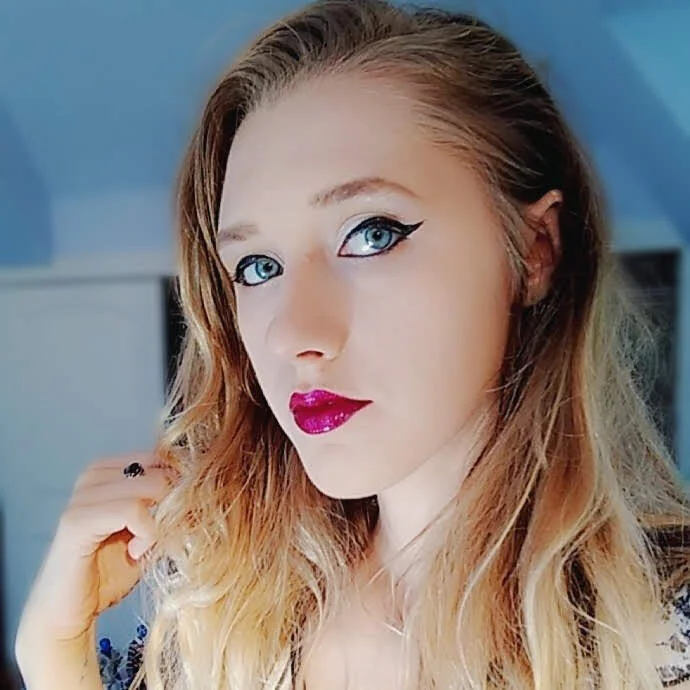A woman with long wavy blonde hair, bright blue eyes with dramatic eyeliner, and shiny pink lipstick, looking at the camera.