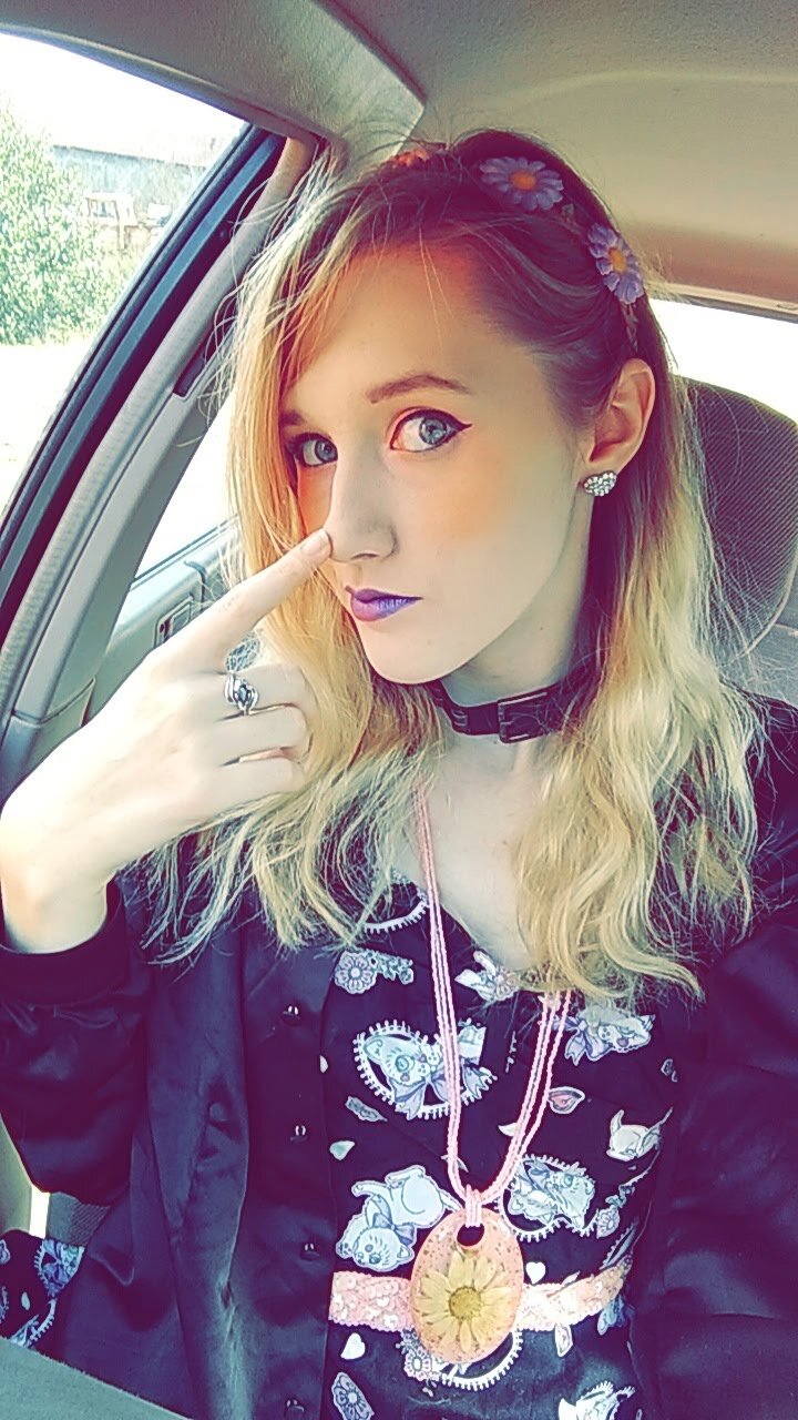 A young woman with blond hair and tattooed arms sitting inside a car, wearing a black shirt with cartoon prints, a choker, floral hairband, earrings, and a pink necklace with a sunflower pendant, touching her nose.