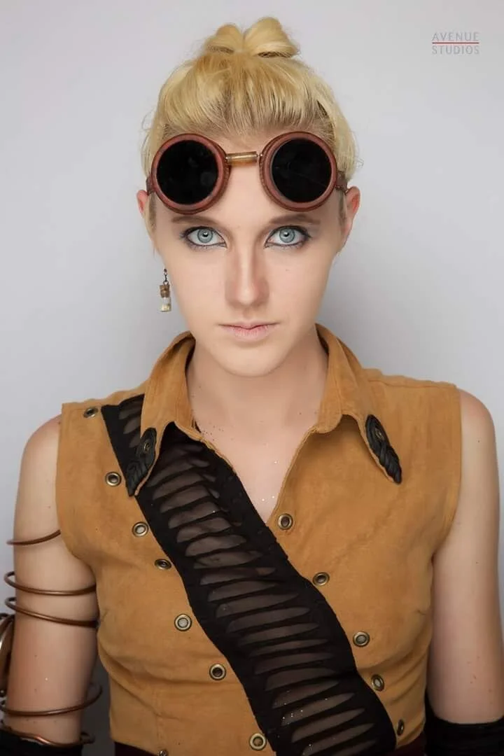 A woman with platinum blonde hair styled in an updo wearing round sunglasses on her forehead, a sleeveless brown top with black mesh detail, silver earrings, and arm cuffs.
