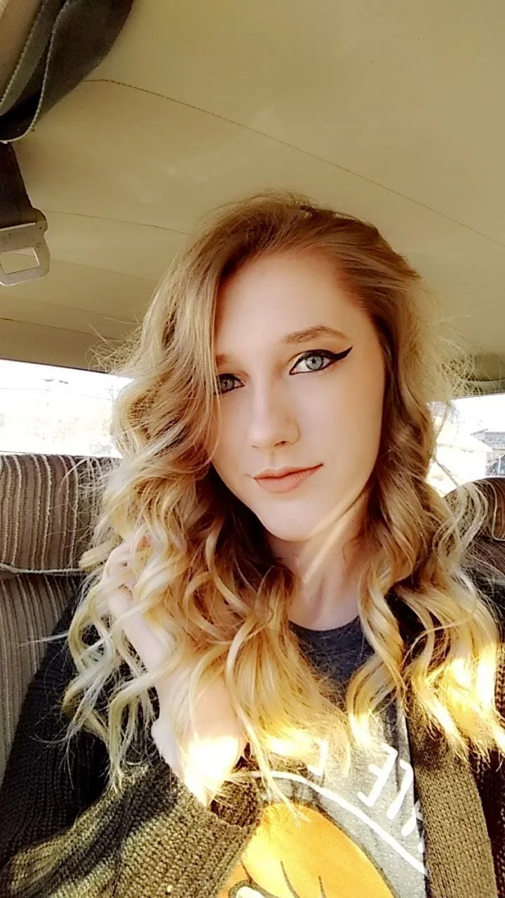 Young woman with blonde, curly hair sitting in a vehicle, wearing a dark top and a cardigan, with winged eyeliner makeup.