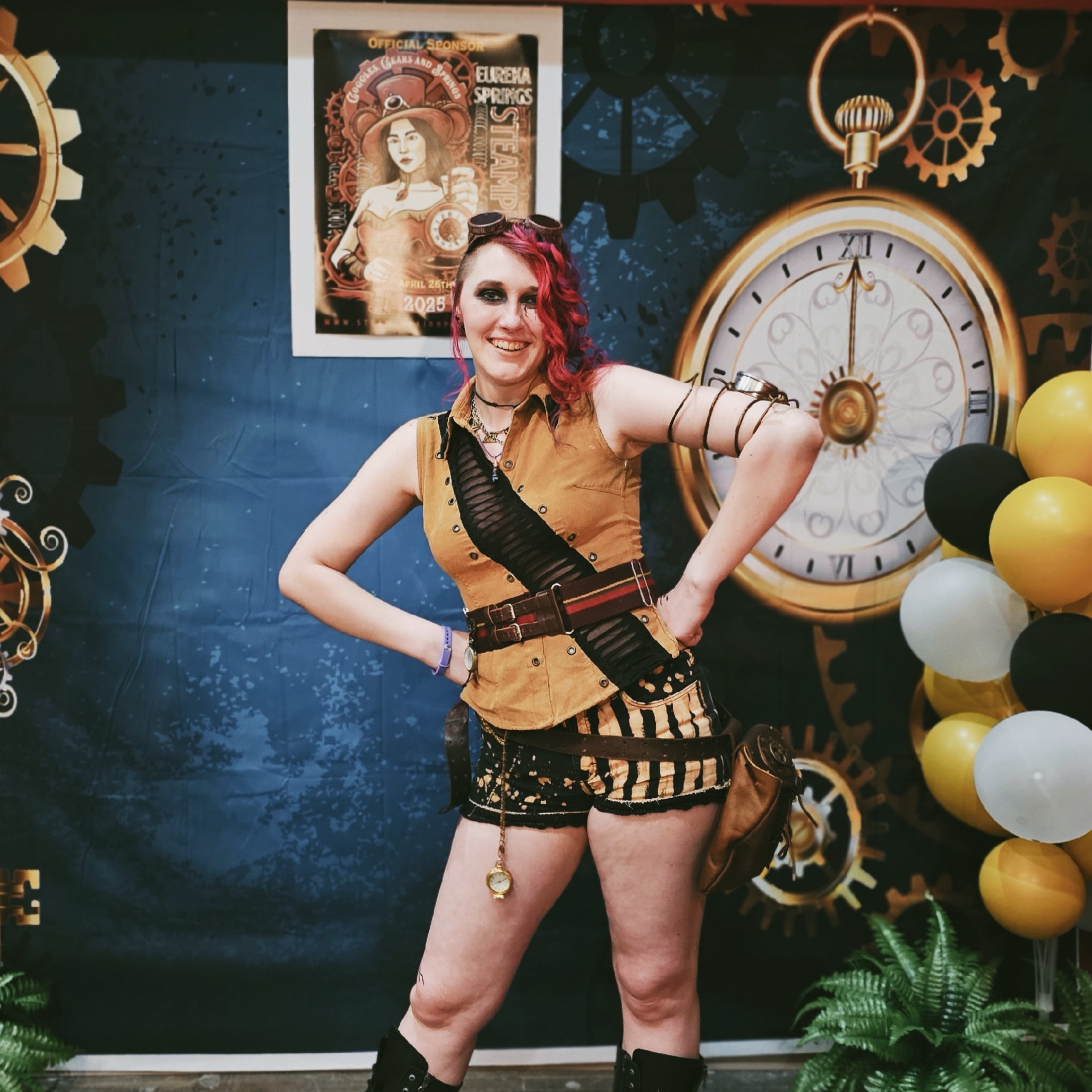 A woman dressed in climbing or punk style clothing with long blonde hair, large round sunglasses, and black platform boots, posing with a vintage rifle while sitting against a large tree trunk and leaning on a textured brown wall.