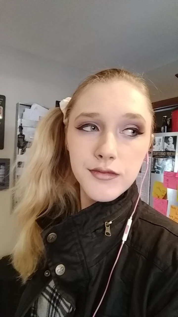 A young woman with blonde wavy hair, wearing a black leather jacket, looking to the side, in an indoor setting with papers and posters on the wall behind her.
