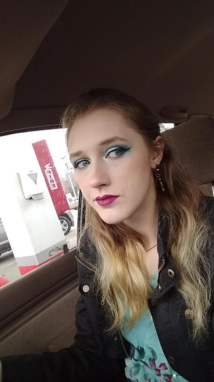 A young woman with makeup sitting in a car, with a visible drive-thru ATM or bank machine behind her.