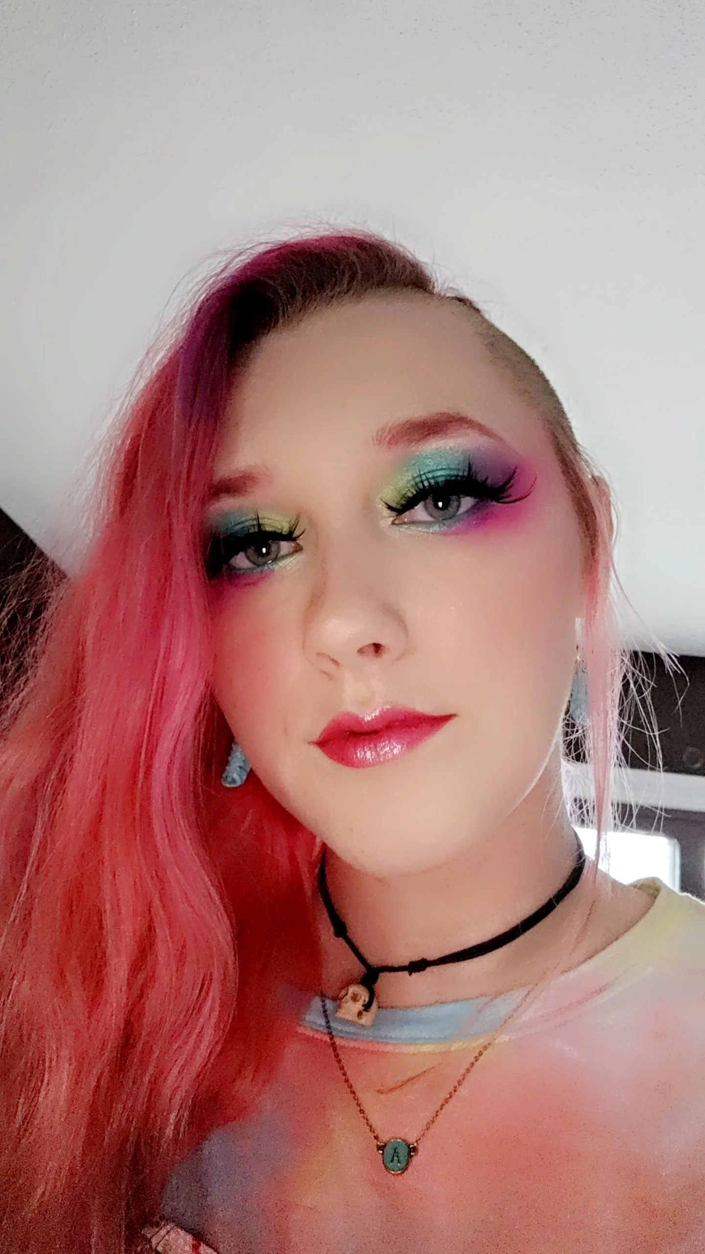 Close-up of a young woman with colorful rainbow eyeshadow, pink hair, and glossy lips, wearing layered necklaces and earrings.