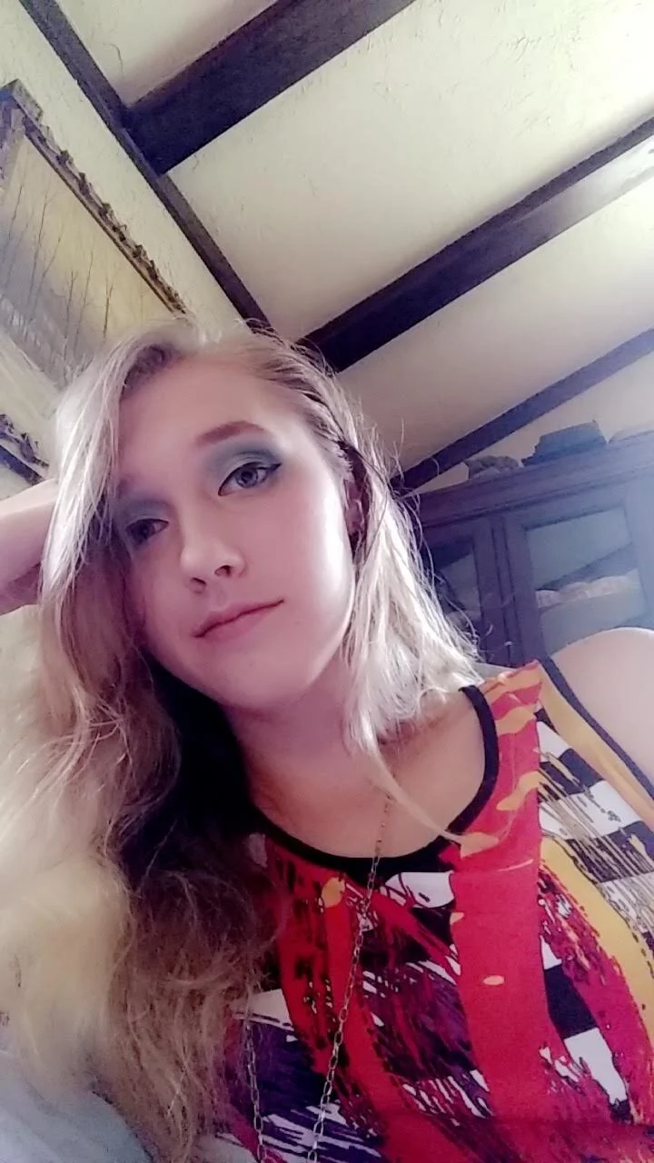 A young woman with long blonde hair and blue eyes, wearing a colorful sleeveless top and a gold chain necklace, sitting indoors against a backdrop of a wooden ceiling and a cabinet.
