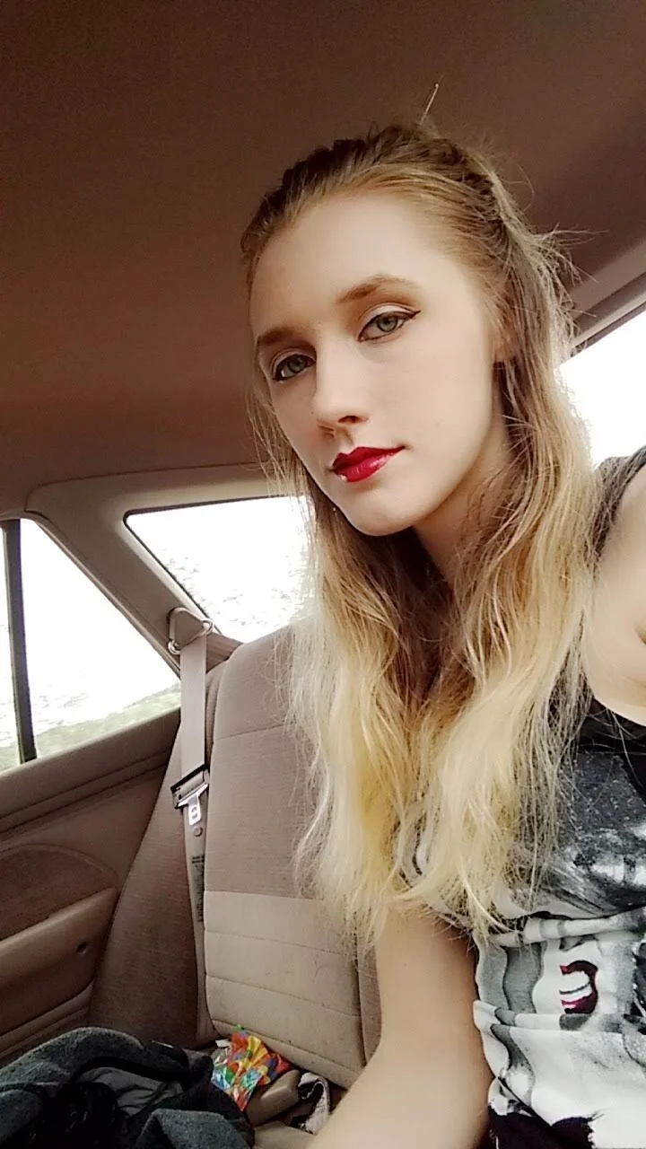 A young woman with long blonde hair and red lipstick taking a selfie in the back seat of a car.