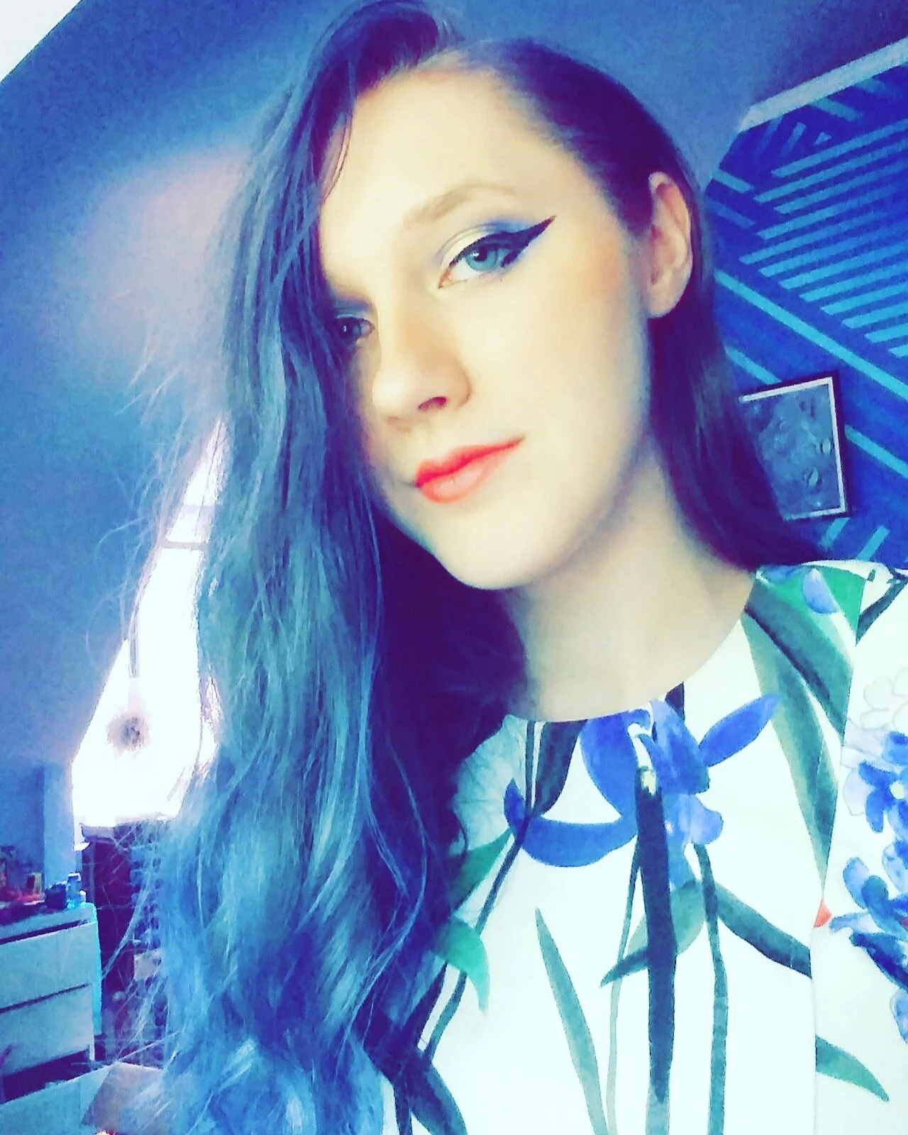 A young woman with long wavy dark hair, blue eyes with winged eyeliner, and coral lipstick, wearing a white top with a blue and green floral pattern, taking a selfie in a room with a dark blue striped ceiling and a framed picture on the wall.
