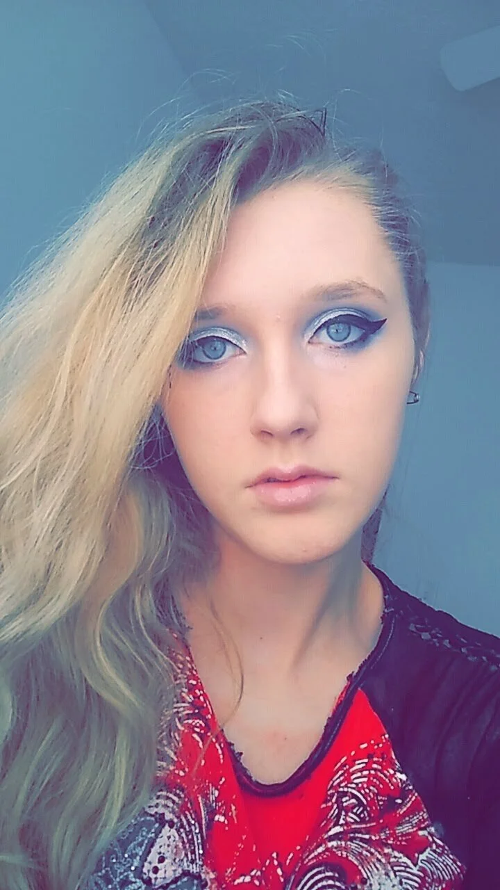 A young woman with long blonde hair and blue eyes, wearing eye makeup with winged eyeliner and eyeshadow, and a red and black patterned shirt.