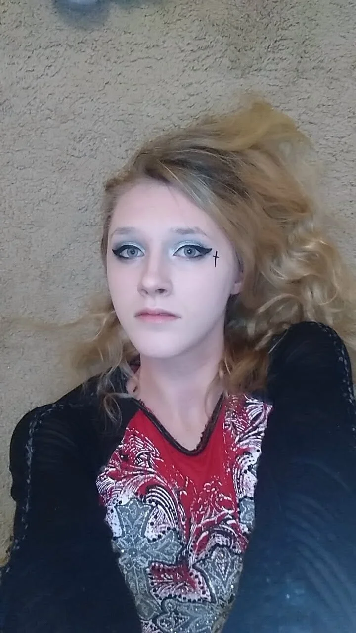 A woman with blonde, curly hair lying on a beige carpeted floor, wearing a red top with a black and white lace pattern, and black long sleeves. She has makeup with bold eyeliner and a small cross tattoo near her right eye.