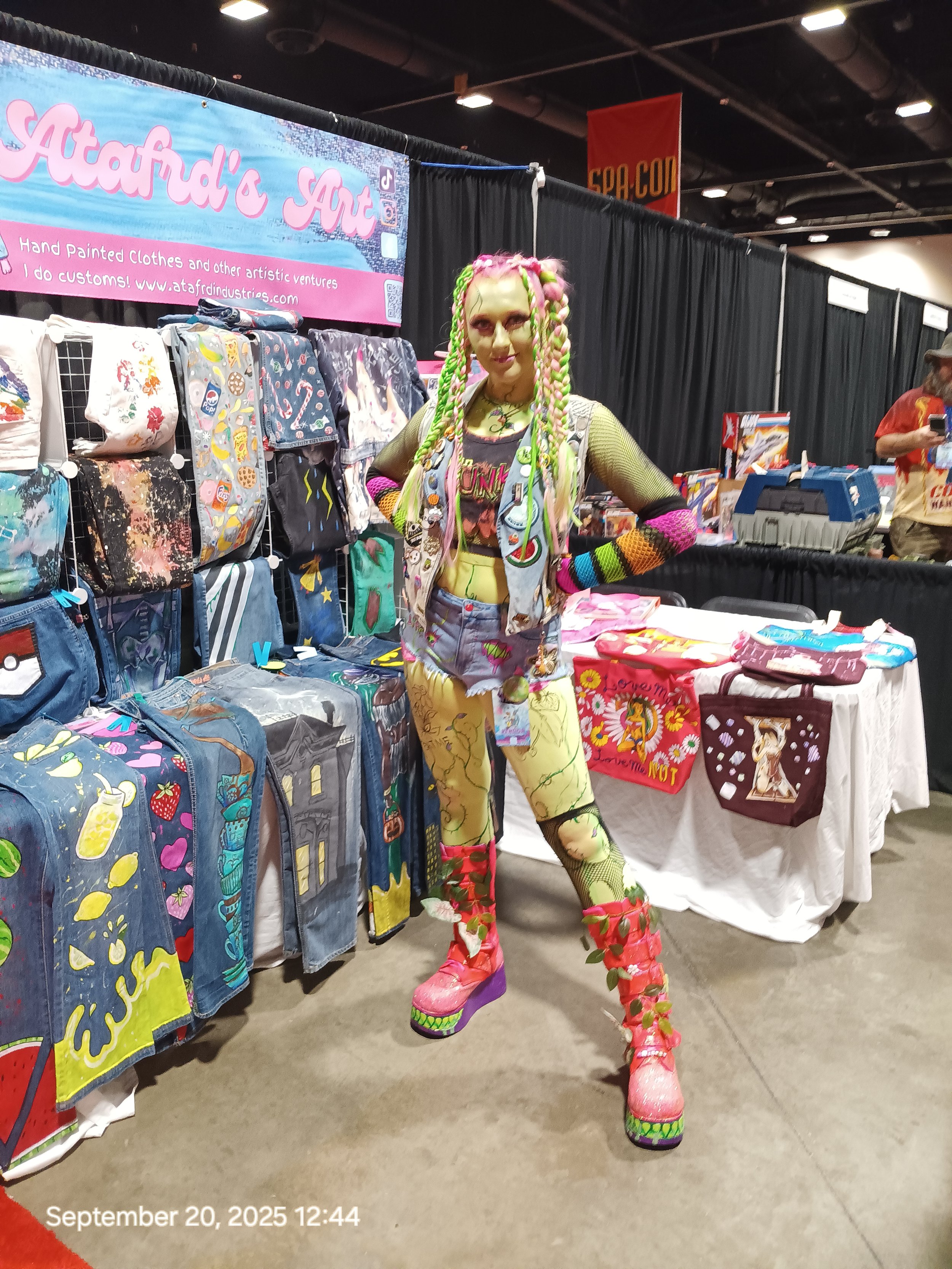 A woman standing in front of a booth at a convention, displaying painted denim jackets and patches. She is wearing a black crop top, striped pants, and pink-heeled shoes. The booth features a pink banner that reads "Atafad's Art" and a display with colorful stickers and buttons.