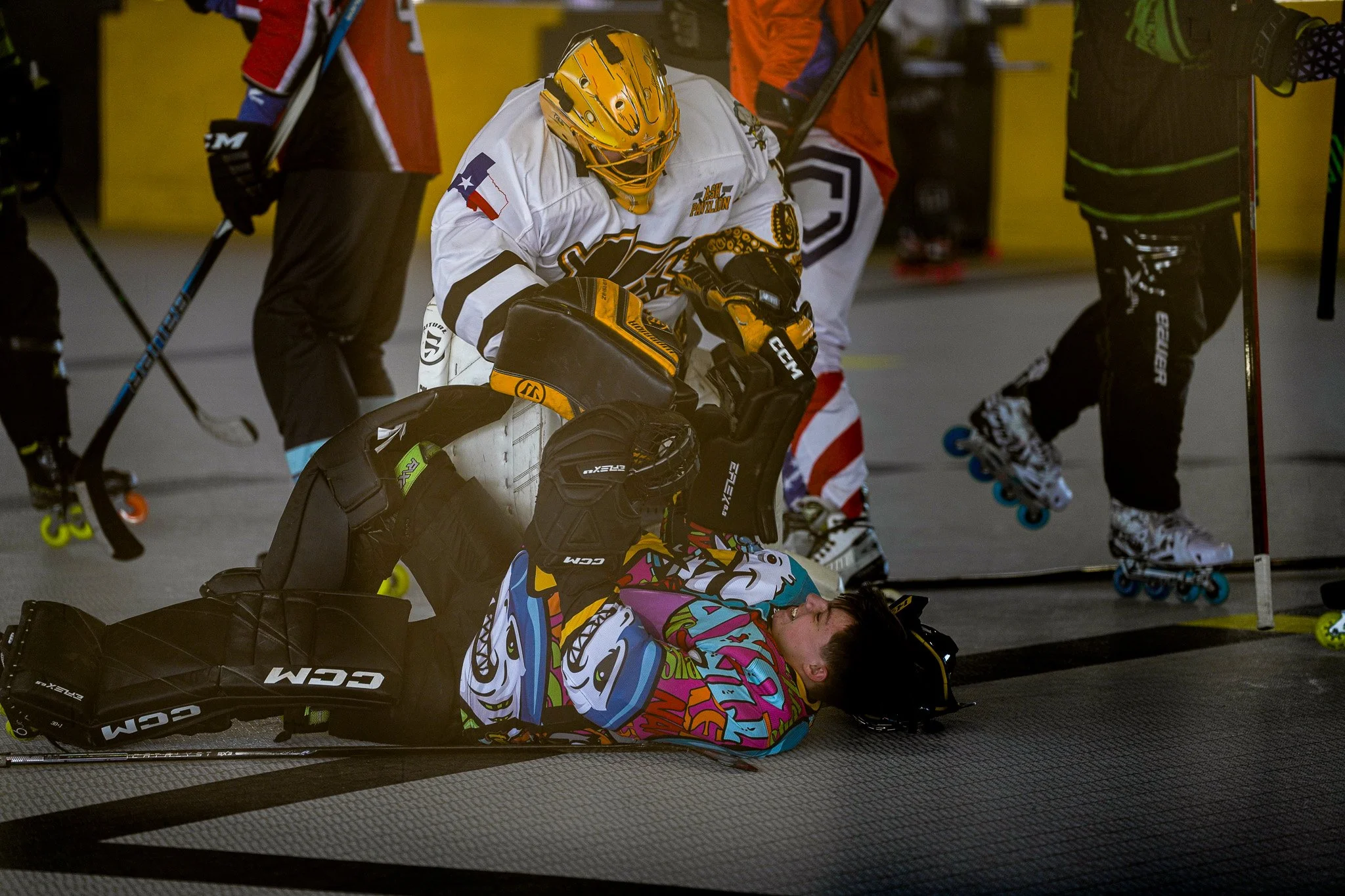 A roller hockey player lying on the floor and being attended to by a teammate while other players stand around on the rink.