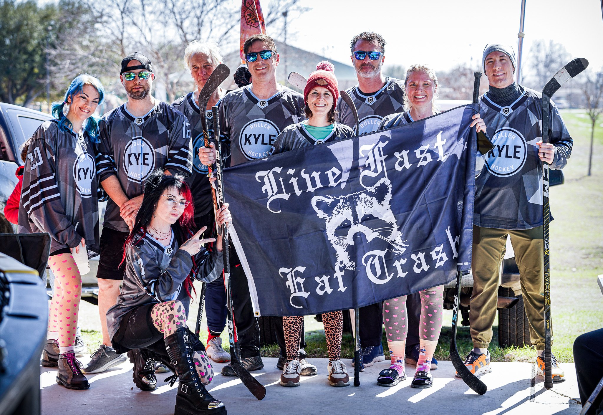 A group of people dressed in hockey jerseys holding a banner that reads 'Live Fast, Eat Trash' with a raccoon illustration. They are outdoors on a sunny day.