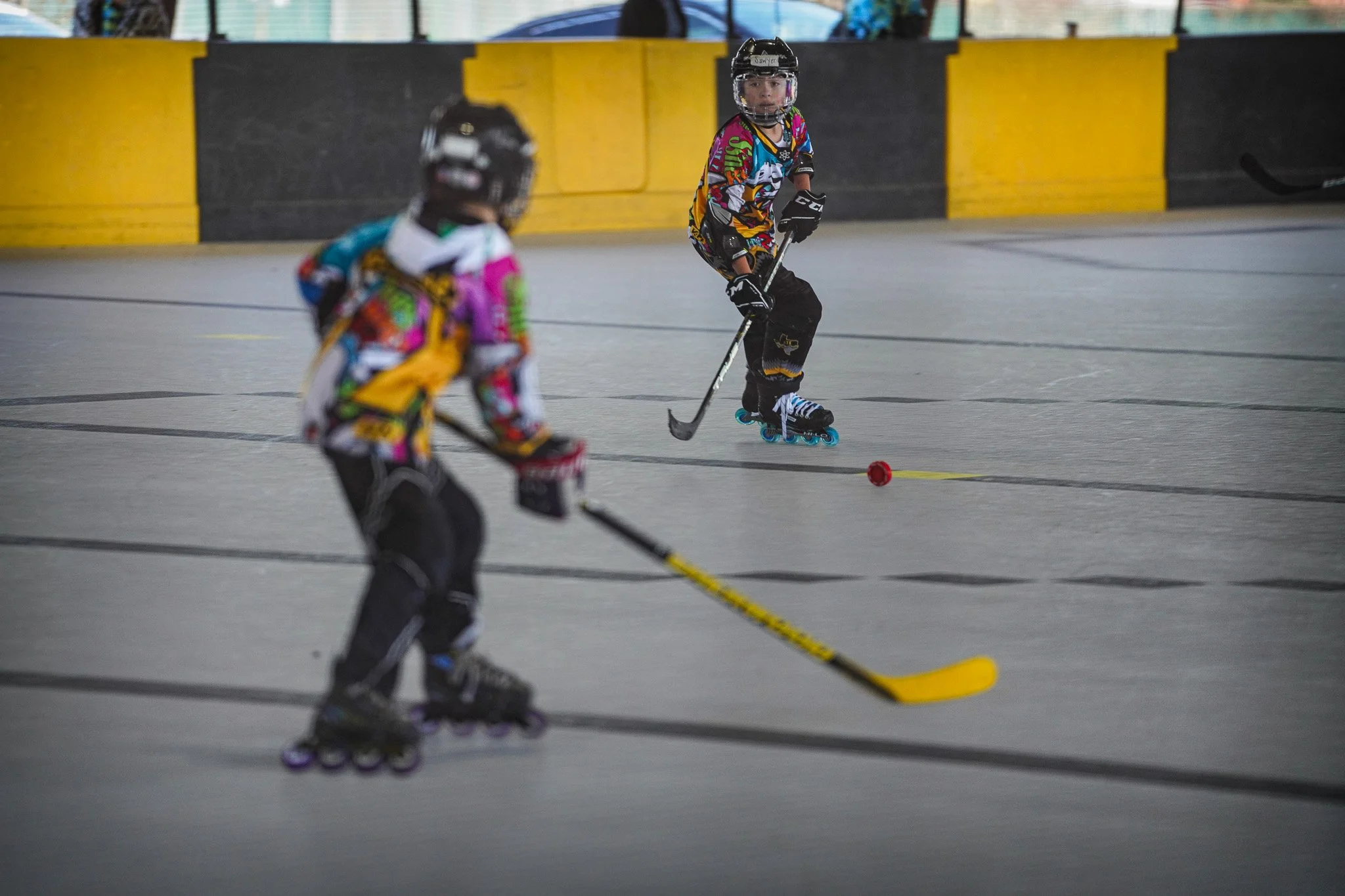 Two children playing roller hockey indoors, wearing colorful jerseys and helmets, with one child in the foreground holding a yellow hockey stick and the other in the background on roller skates, near a hockey puck.