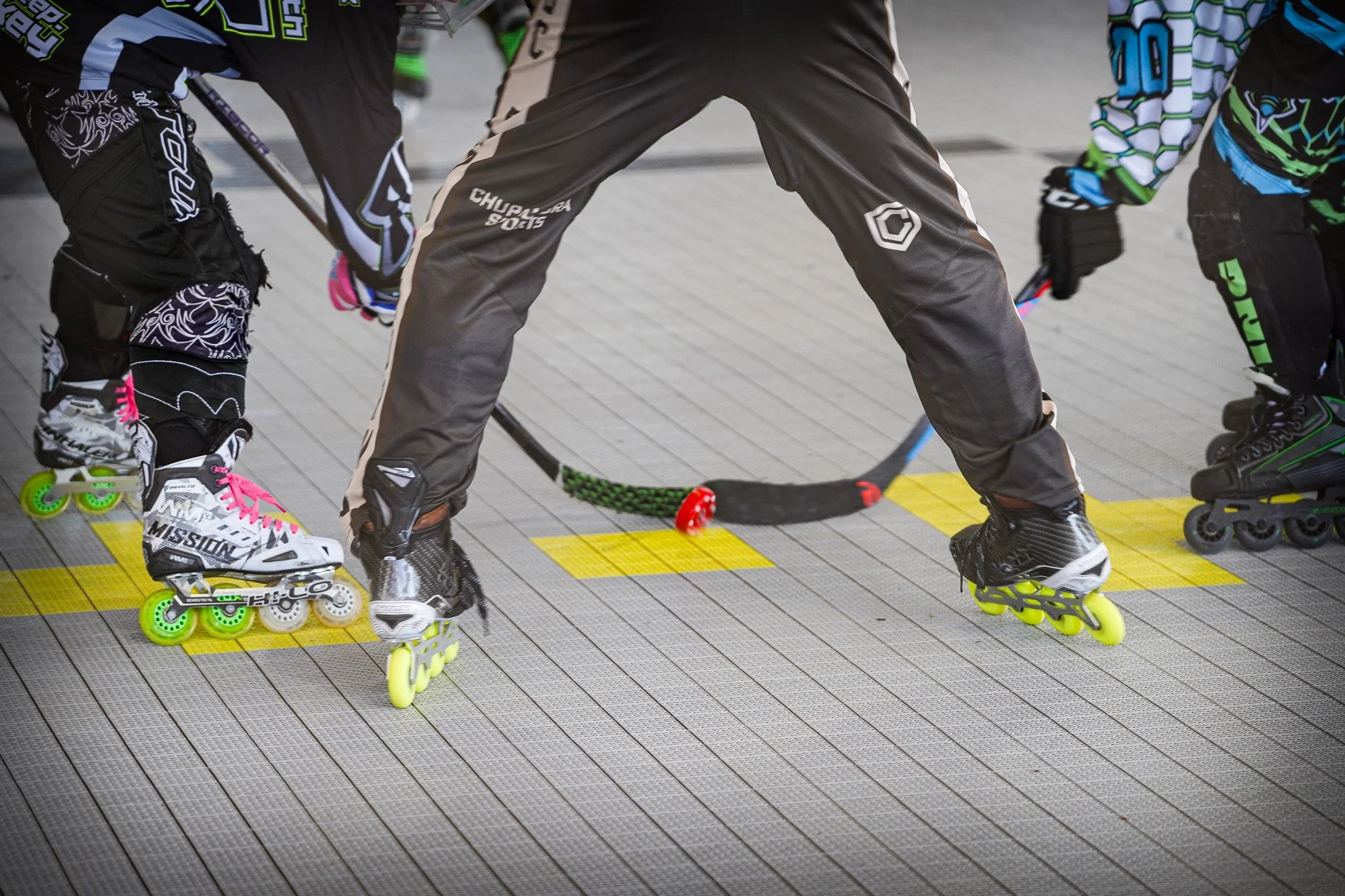 Close-up of children and a coach playing street hockey on rollerblades, with a focus on their legs and hockey sticks, on a gray surface marked with yellow lines.