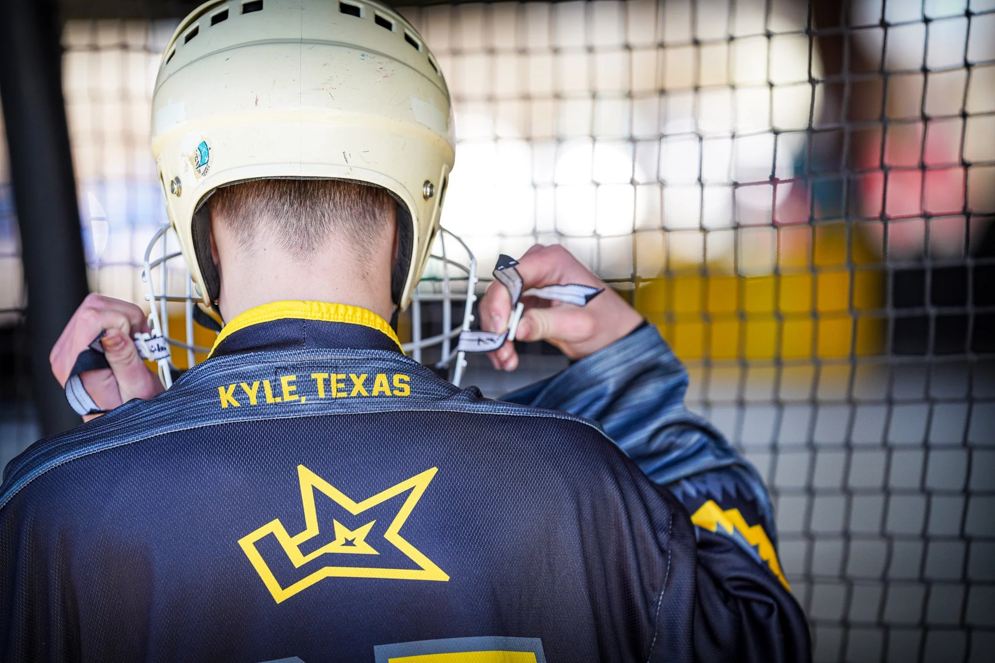 Back of a young baseball player from Kyle, Texas, adjusting a helmet in a dugout, wearing a dark jersey with yellow lettering and a star logo.