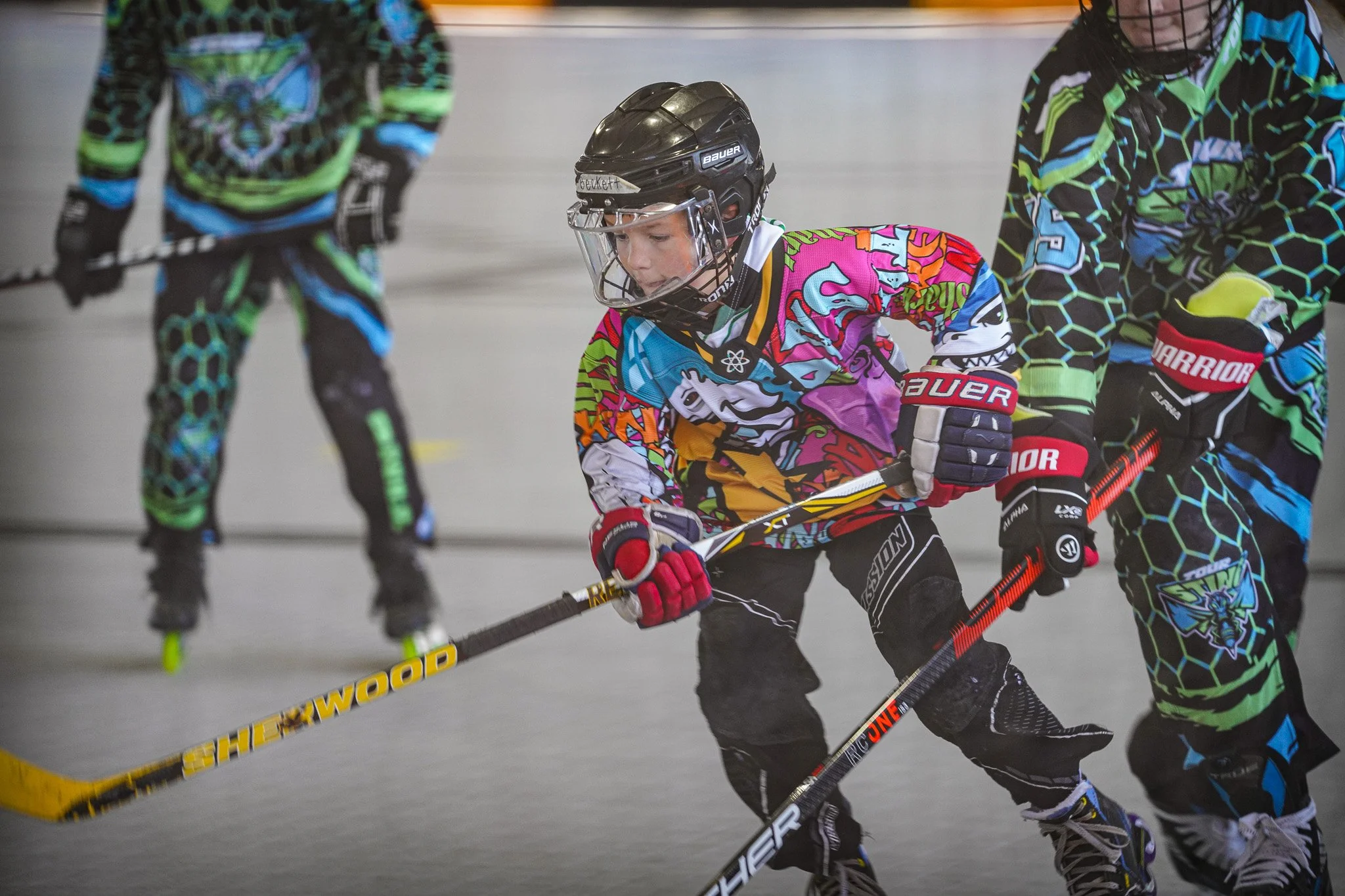 Young boy wearing a colorful hockey jersey, black helmet, and hockey gloves playing roller hockey. He is using a hockey stick to control the puck along with two other players on an indoor roller hockey rink.