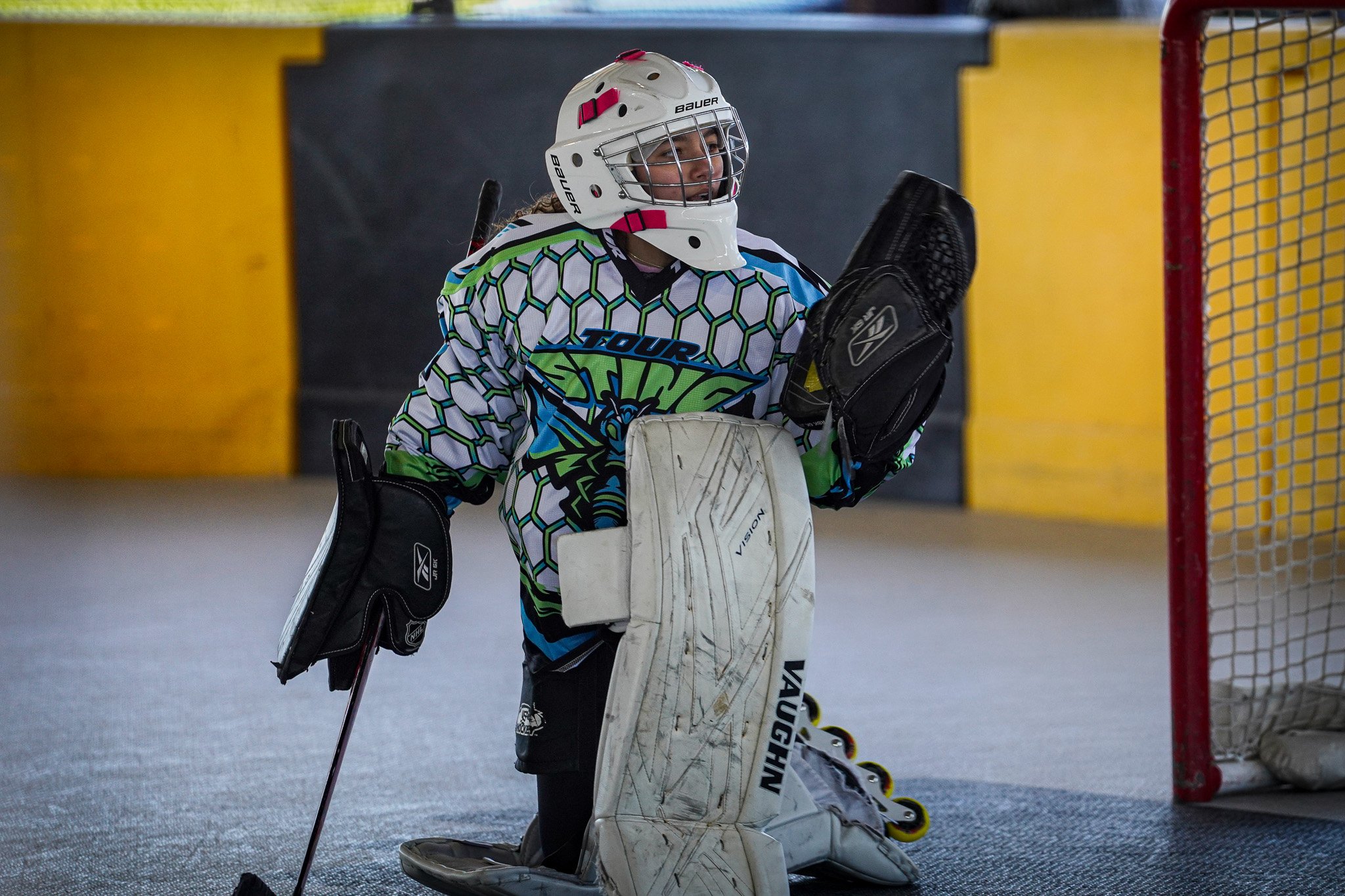 Hockey goalie wearing a colorful jersey, white helmet, and pads, standing near the net in an indoor rink.