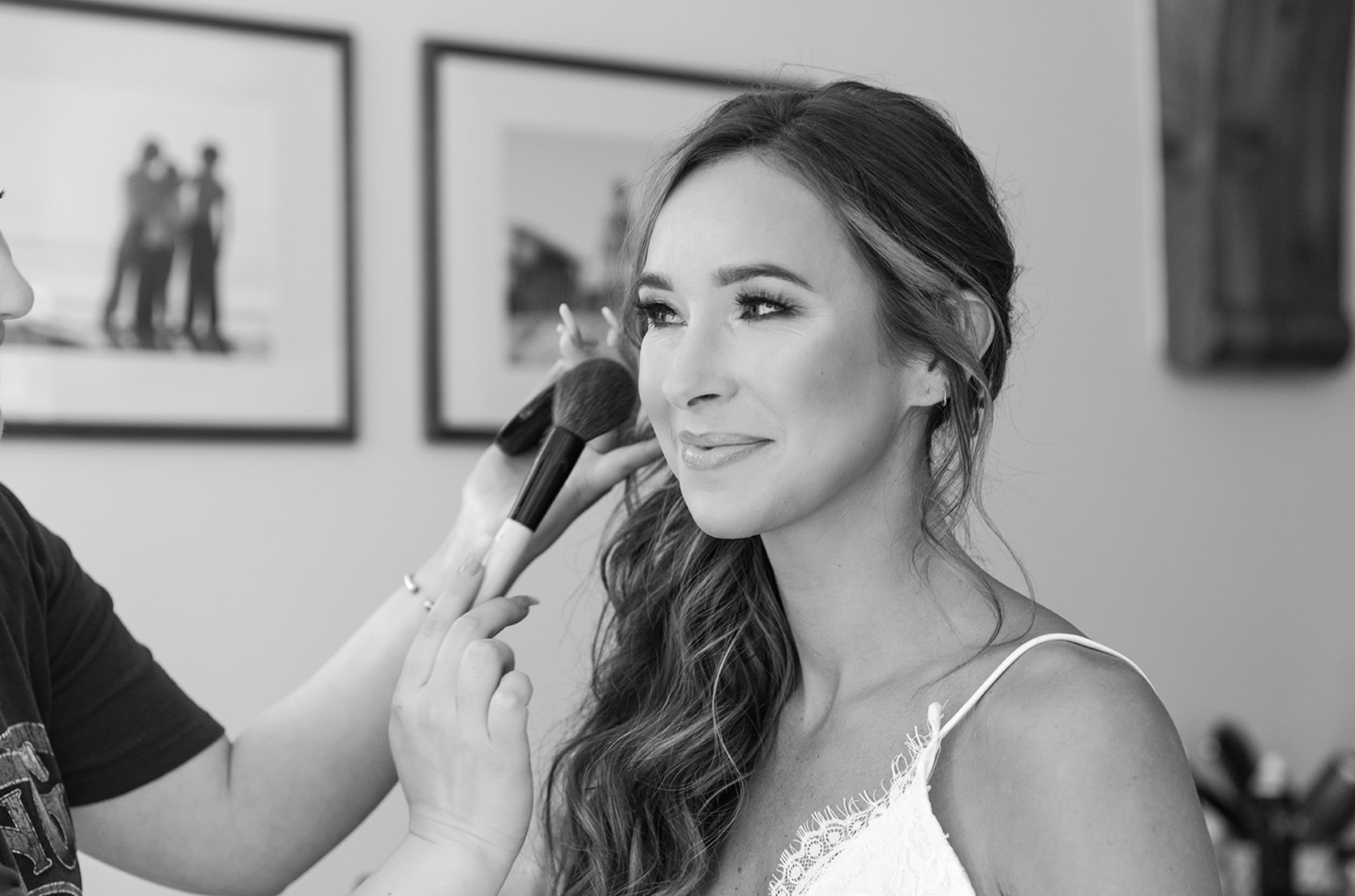 A woman getting her makeup done, with a makeup artist applying blush or highlighter on her cheeks using a makeup brush. She is smiling slightly and looking to the right. The background shows framed photographs on the wall.