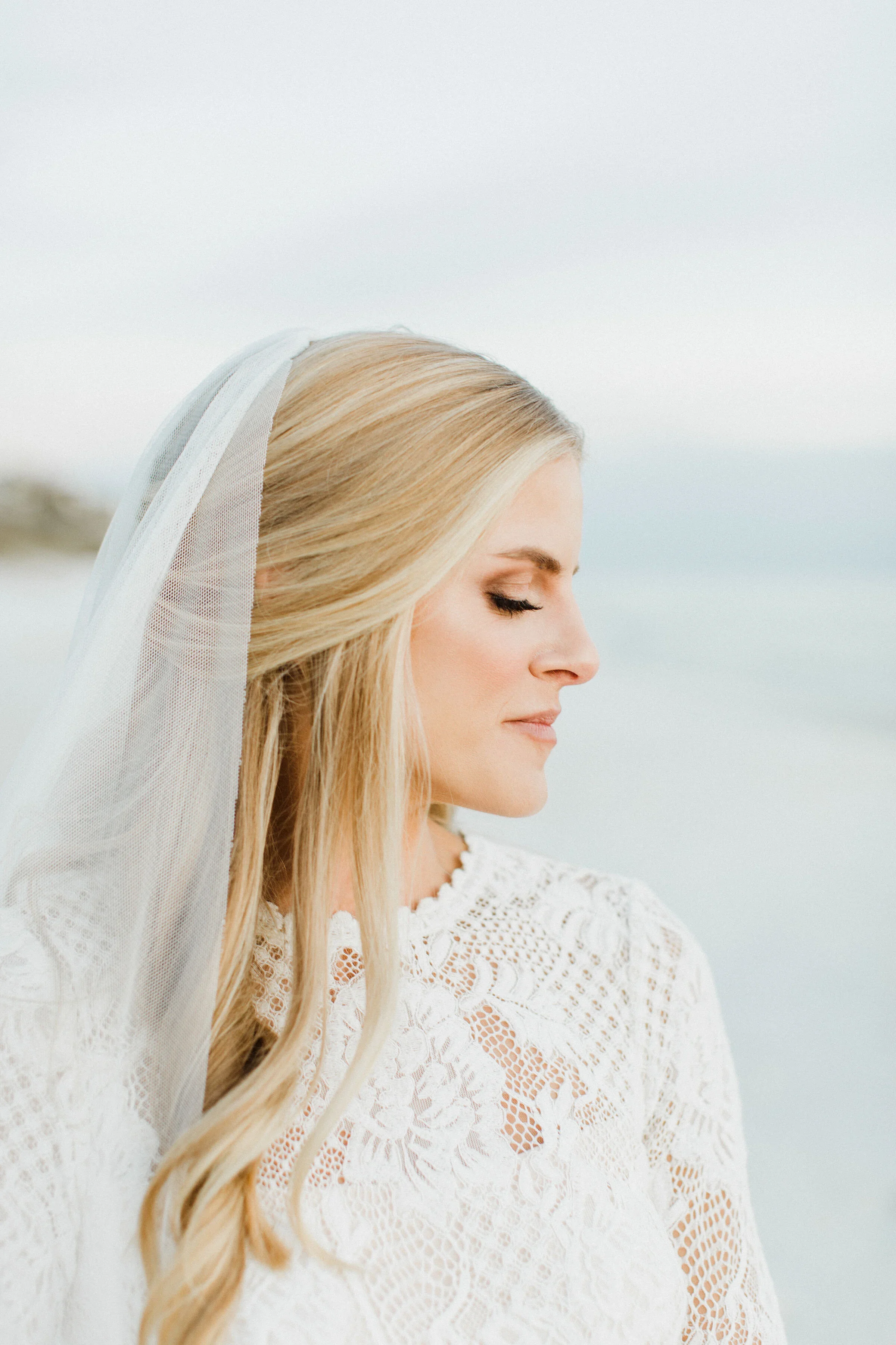 A woman in a white lace dress with a veil, standing outdoors with a body of water and sky in the background.