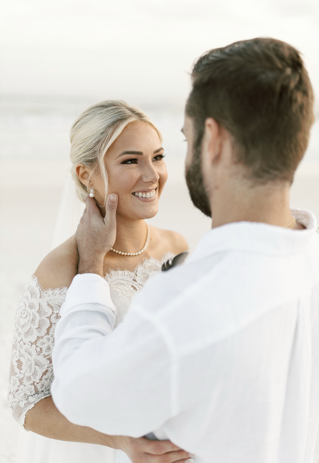 A smiling bride with blonde hair and a pearl necklace looking at her groom on the beach