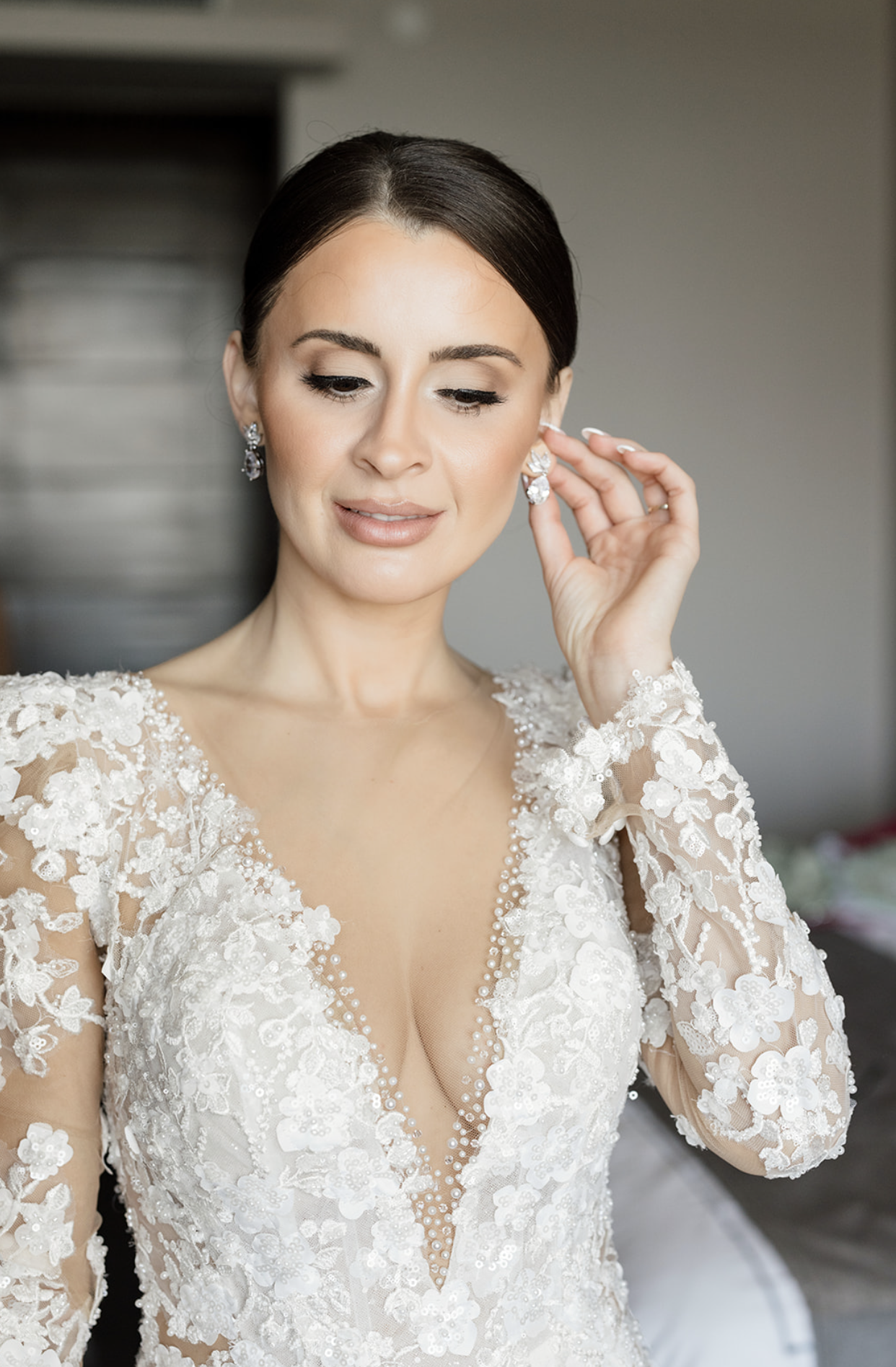 Bridal woman wearing an ornate lace wedding dress with floral embroidery, adjusting her earrings.