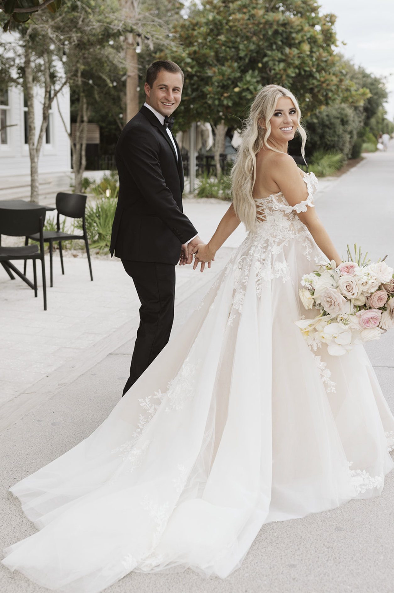 A newlywed couple holding hands outdoors, with the bride in a white wedding gown holding a bouquet of flowers, and the groom in a black tuxedo, smiling and looking at the camera.