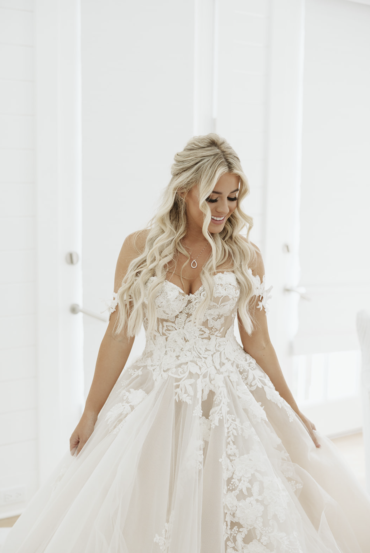 A smiling bride in a white lace wedding gown with off-the-shoulder sleeves.