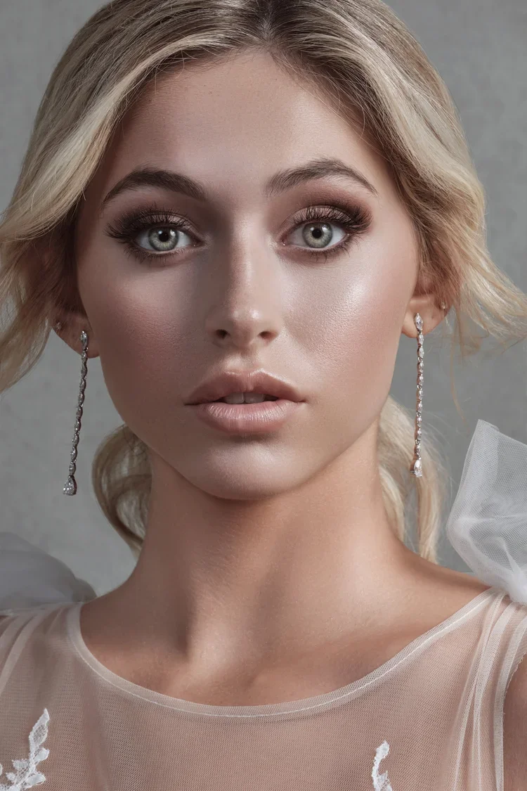 Close-up portrait of a young woman with blonde wavy hair, wearing makeup, dangling earrings, and a sheer white top with embroidery.