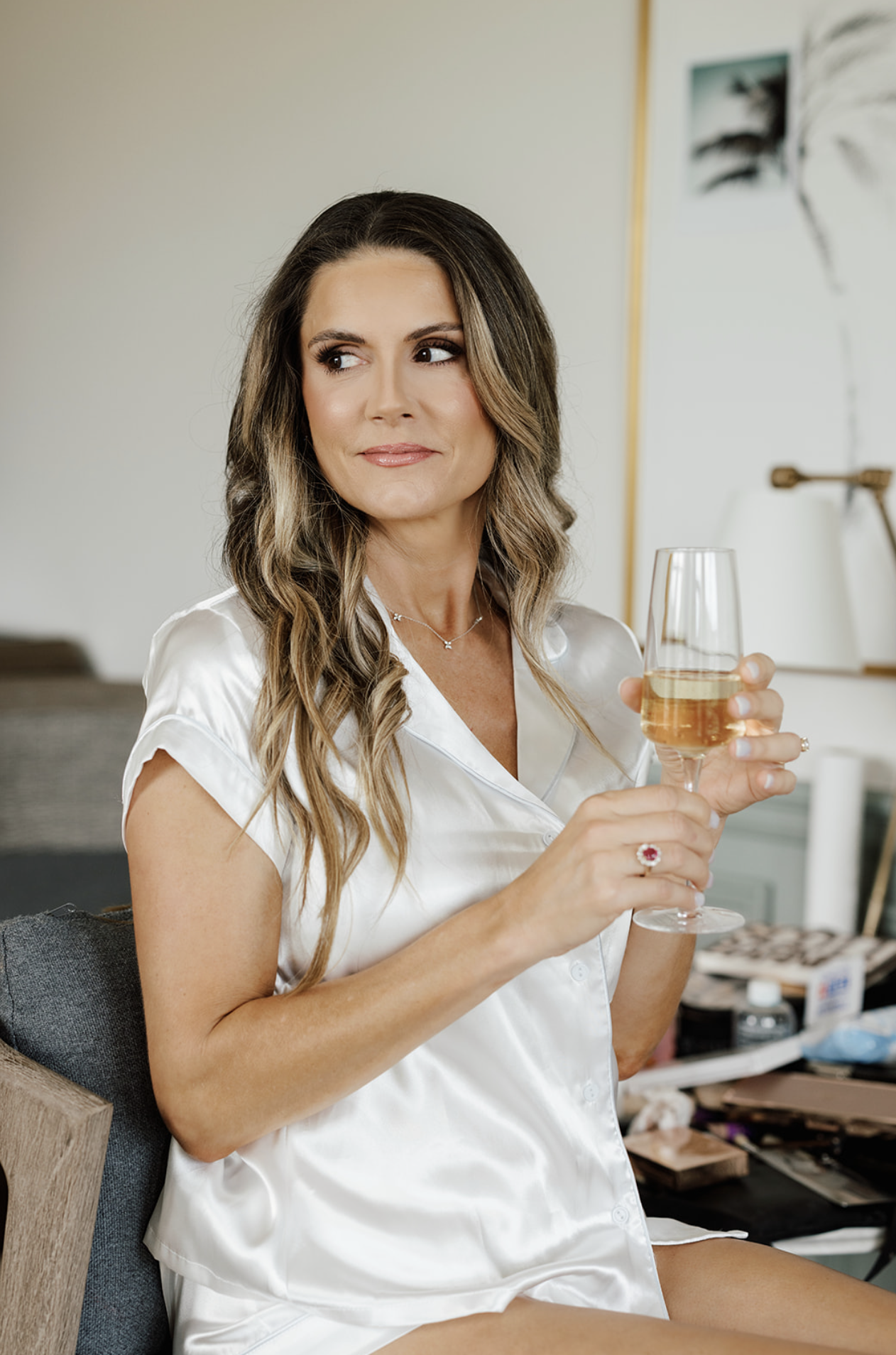 A woman with wavy brown hair wearing a satin white silk pajama top, sitting in a room, holding a glass of white wine, with makeup and jewelry, looking to her left.