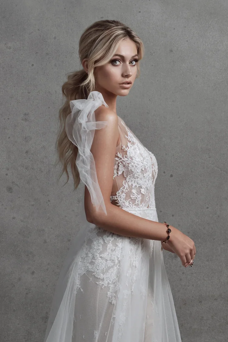 A woman in a white lace wedding dress with tulle shoulder accents, standing against a gray background.