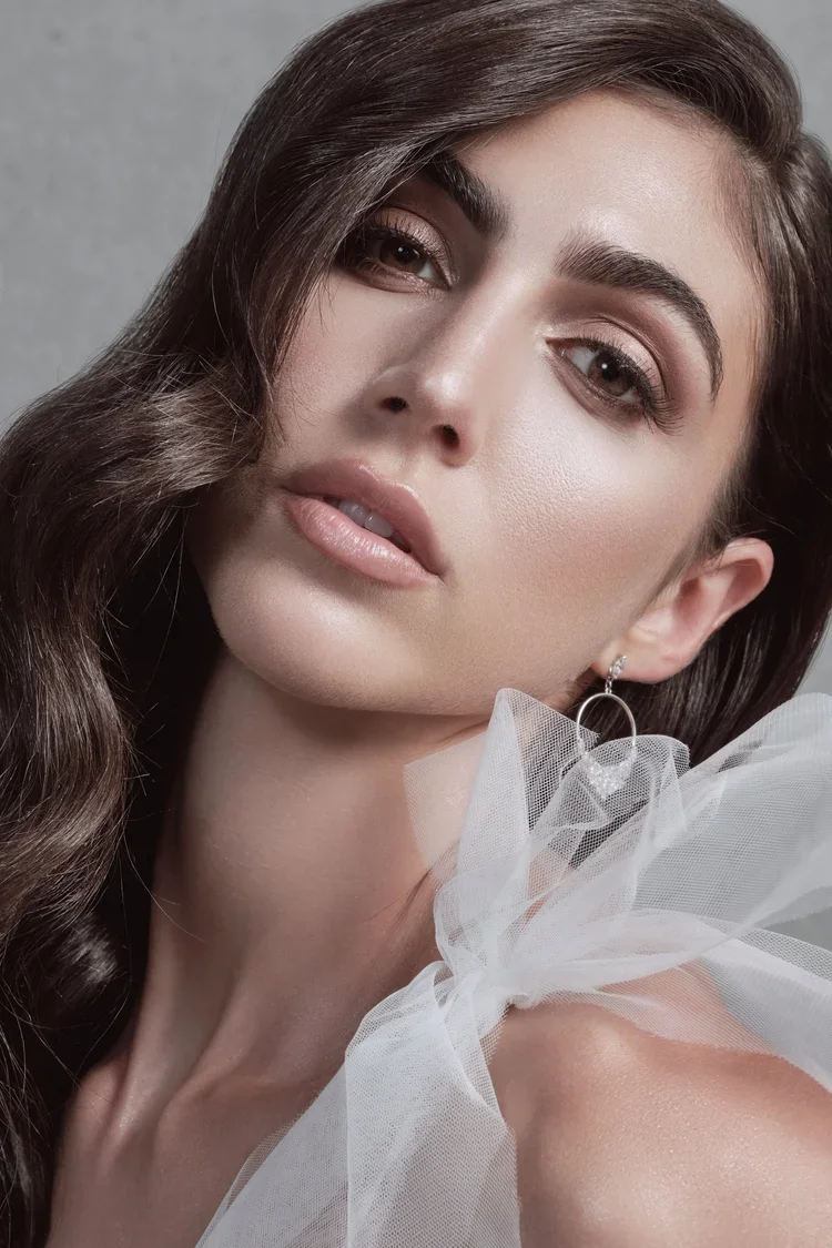 Close-up of a woman with long wavy brown hair and light makeup, wearing a white tulle fabric on her shoulder and a silver earring.