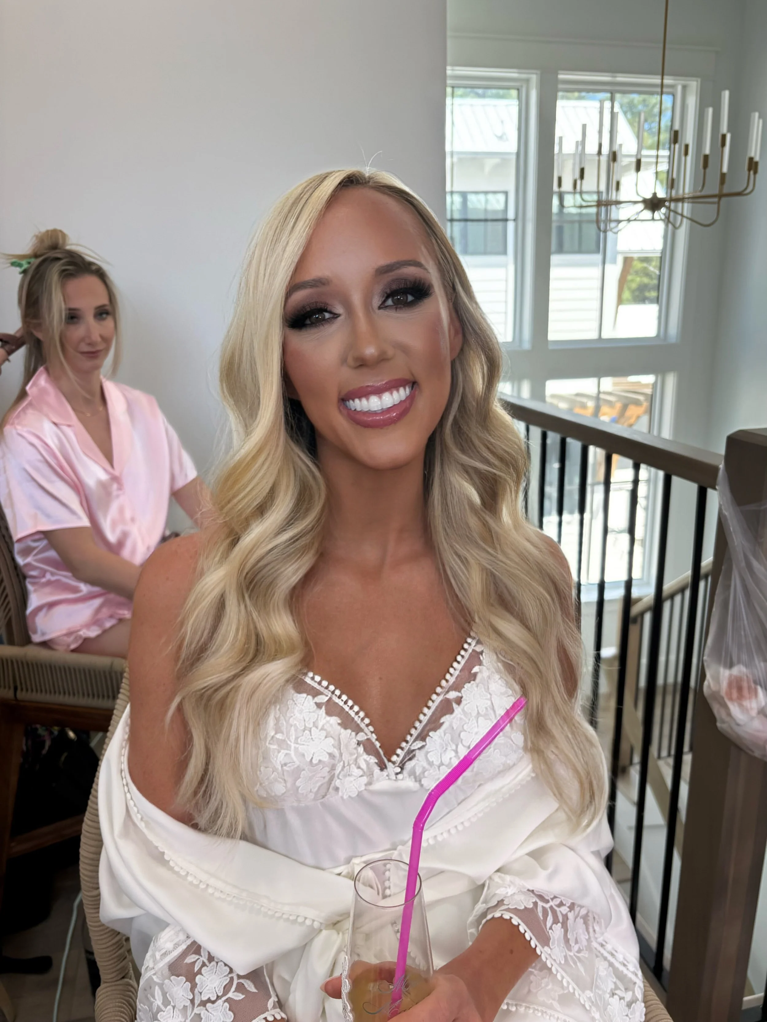 A smiling blonde woman with wavy hair in a white lace dress holding a drink with a pink straw, sitting indoors with another woman in pink satin robe in the background.