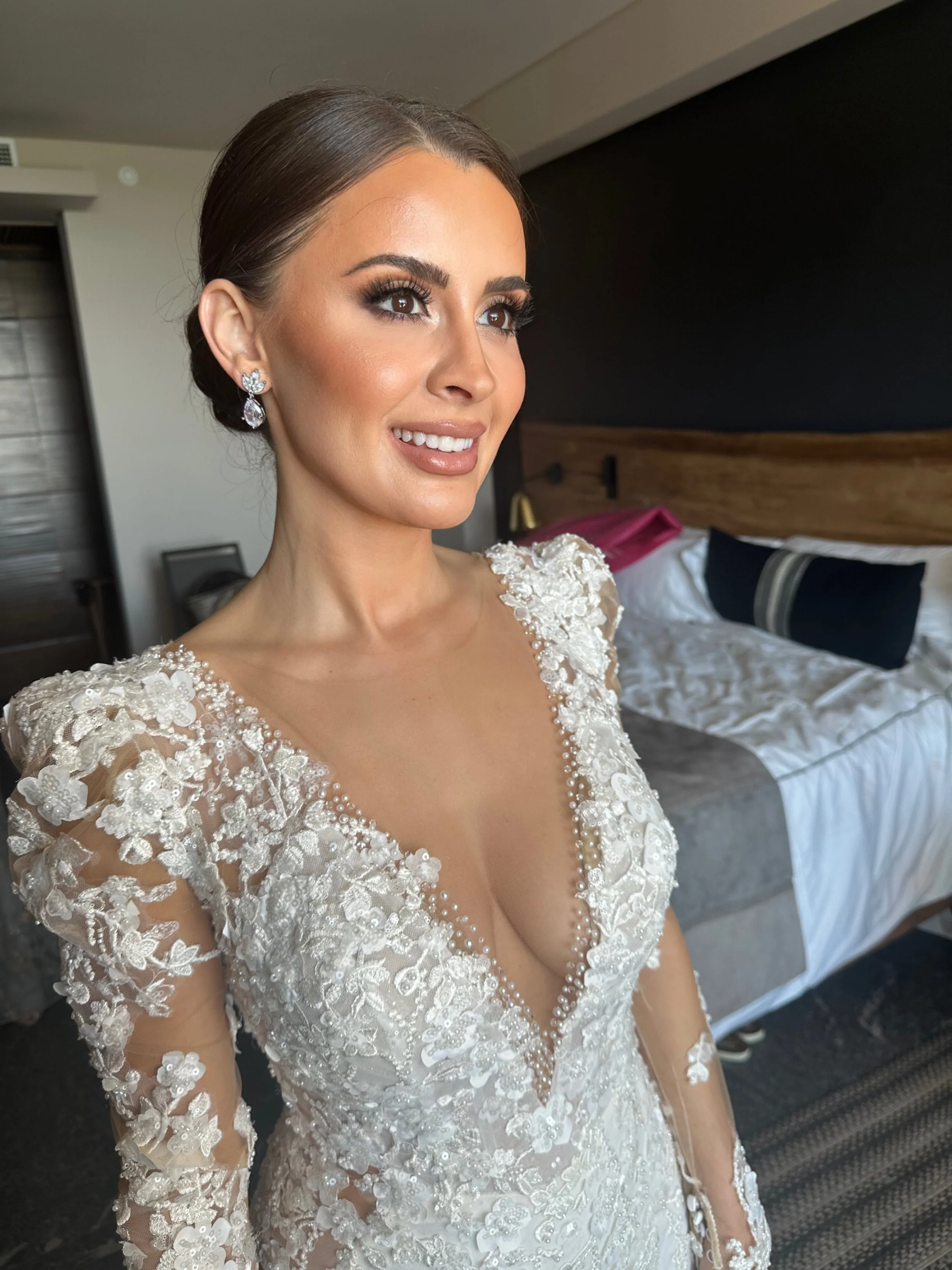 A bride in a white lace wedding dress with floral embroidery, pearl details, and sheer long sleeves, smiling in a hotel room.