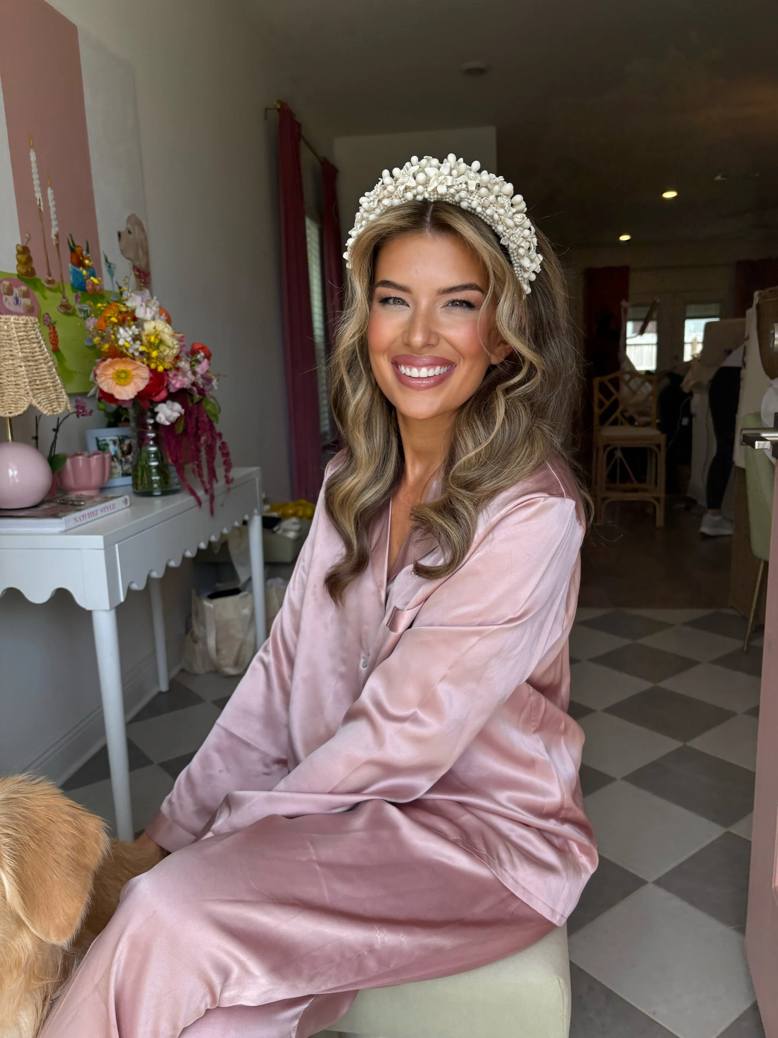 A young woman with long wavy hair and a floral headpiece, wearing pink satin pajamas, smiling and sitting on a chair in a decorated room with flowers and art on the wall.