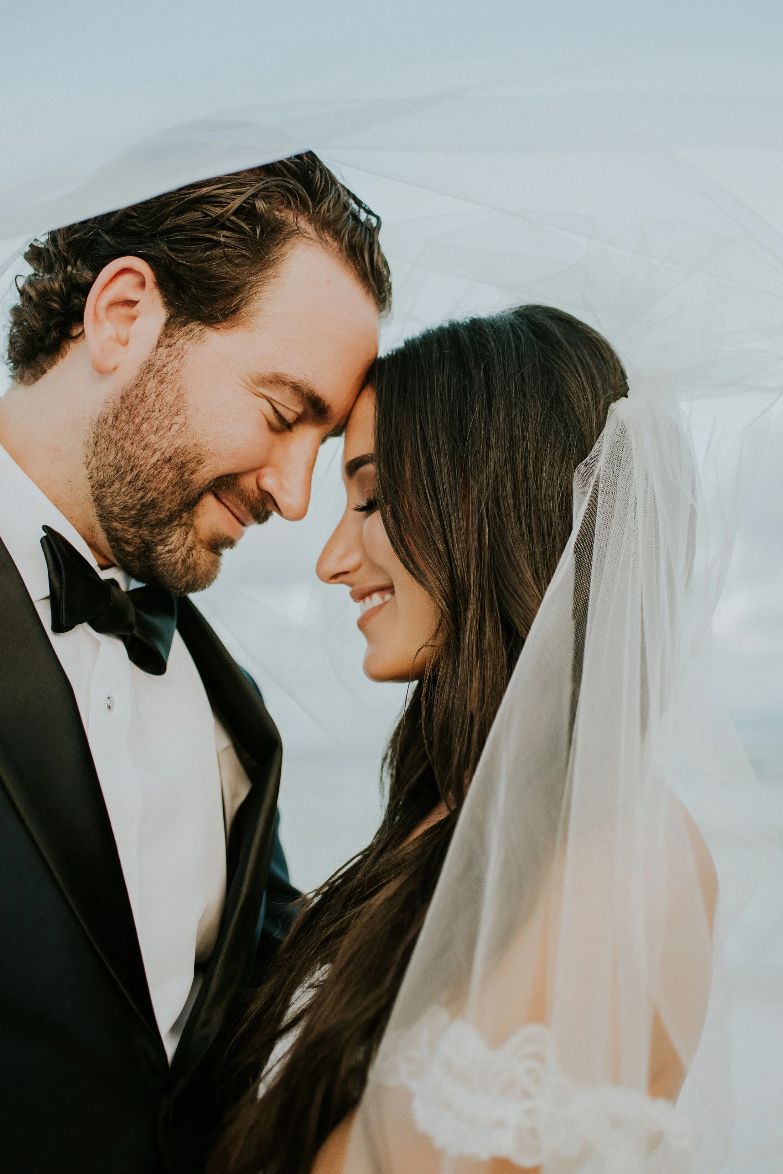 A close-up of a bride and groom with their foreheads touching, smiling, under a white veil, dressed in wedding attire.