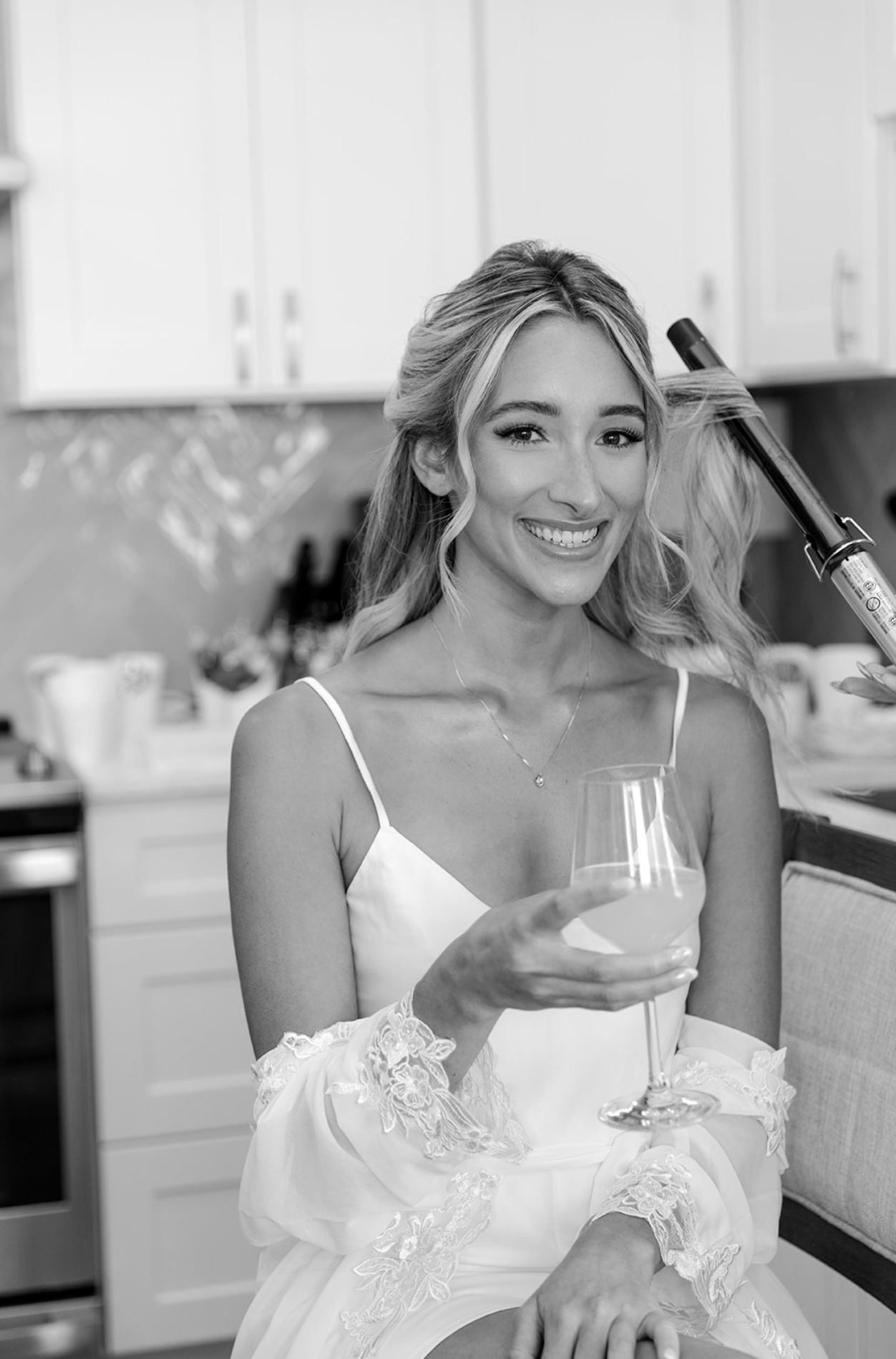 A woman in a white dress smiling while holding a wine glass in a kitchen, a curling iron being used on her hair.