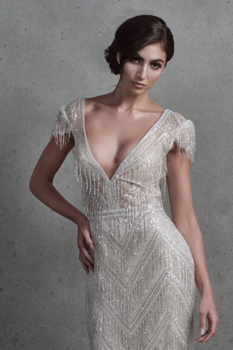 A woman with short dark hair wearing a glamorous, silver, beaded gown with a deep V neckline and fringe detailing on the shoulders, standing against a gray background.
