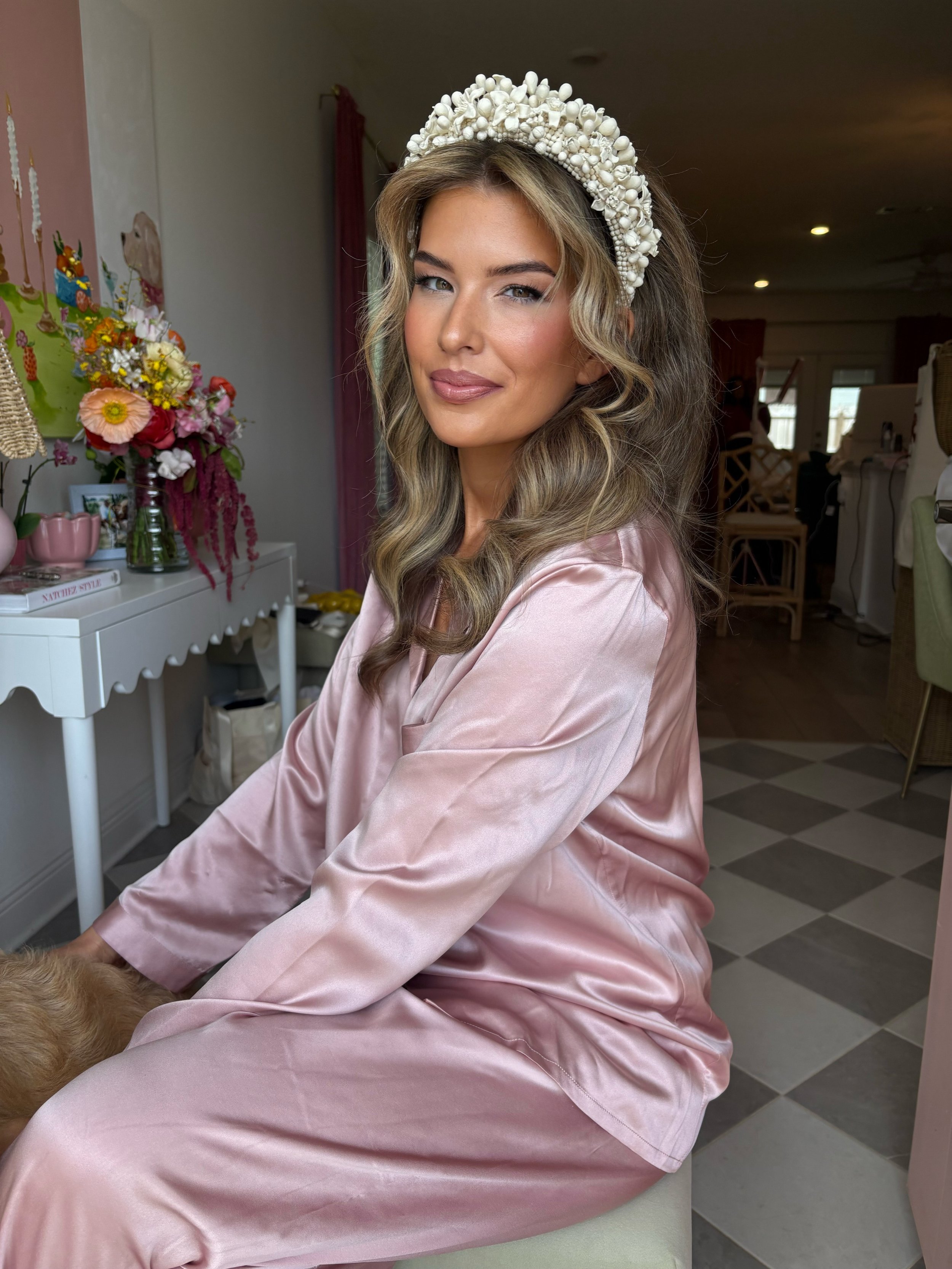 A woman with wavy blonde hair wearing a pearl beaded headband and pink satin pajamas, sitting indoors with a flower arrangement and home decor in the background.