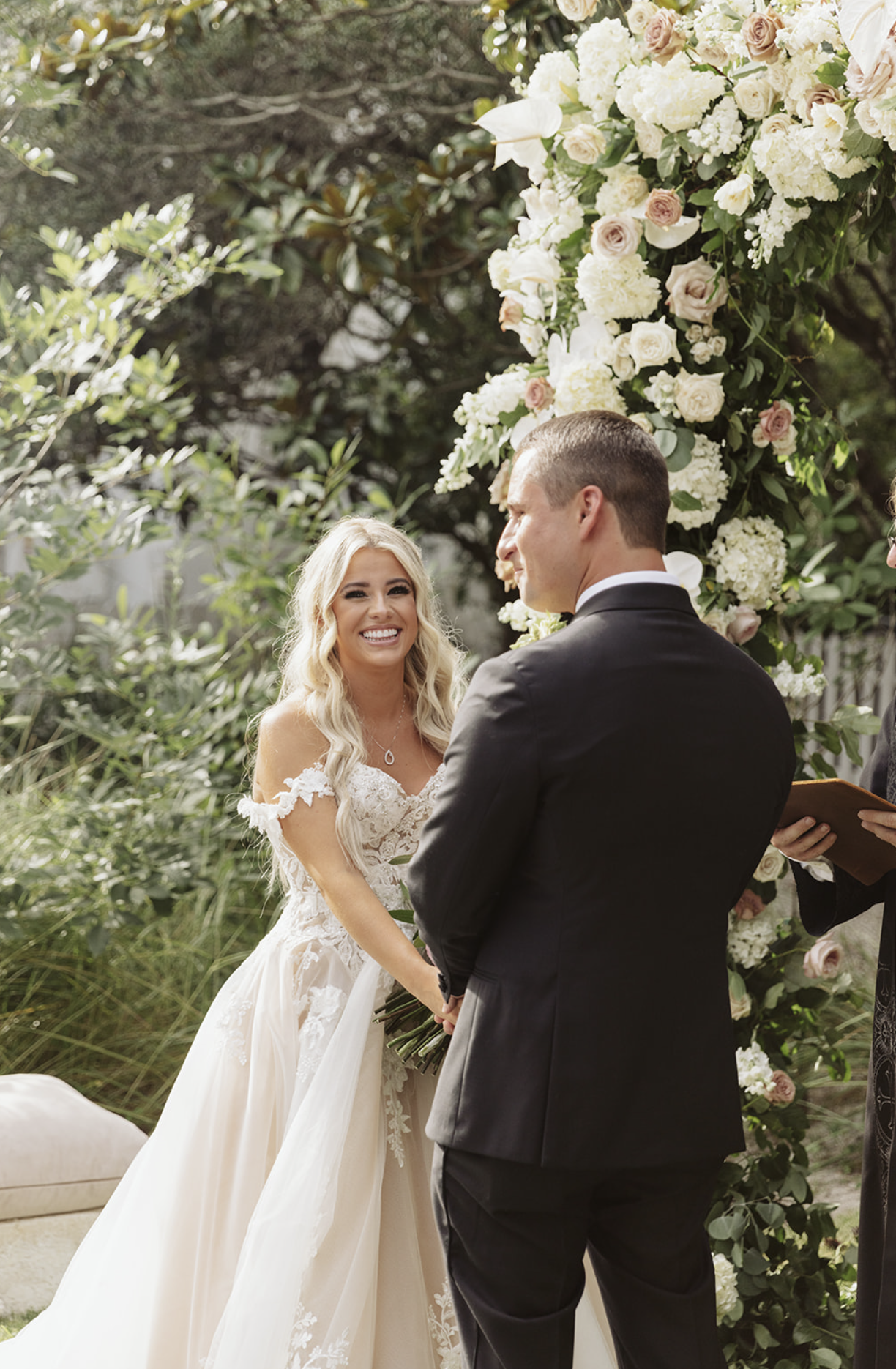 A bride and groom exchanging vows outdoors during a wedding ceremony, with a floral arch behind them and lush greenery surrounding them.