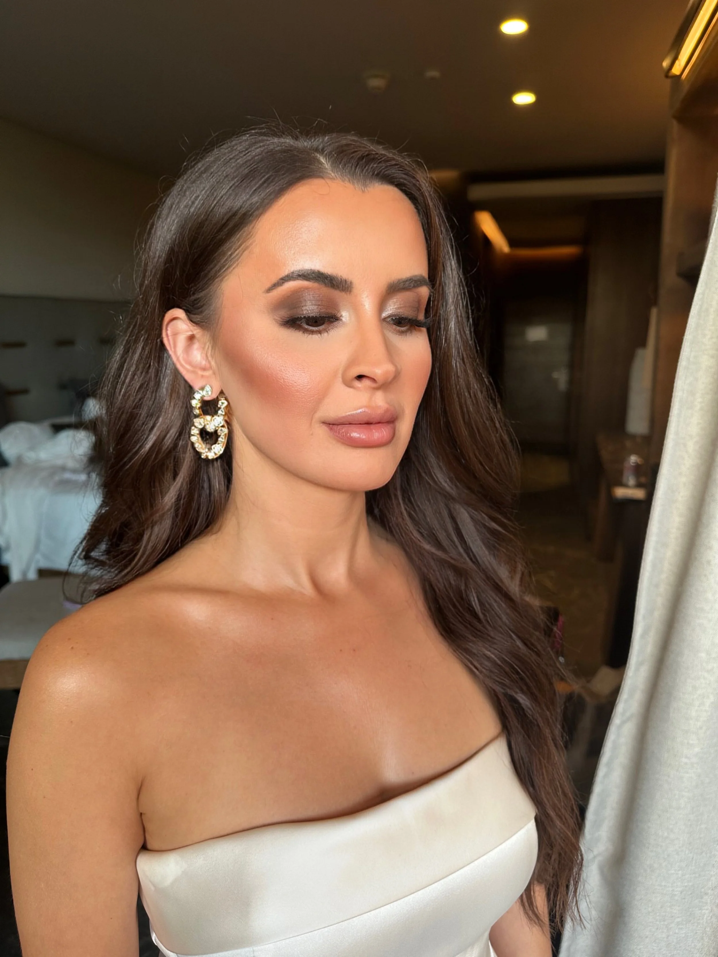 A woman with long brown hair styled in loose waves, wearing large pearl and gold hoop earrings with heart shapes, makeup with smoky eyeshadow, and a strapless white dress, stands indoors near a curtain in a room with warm lighting.