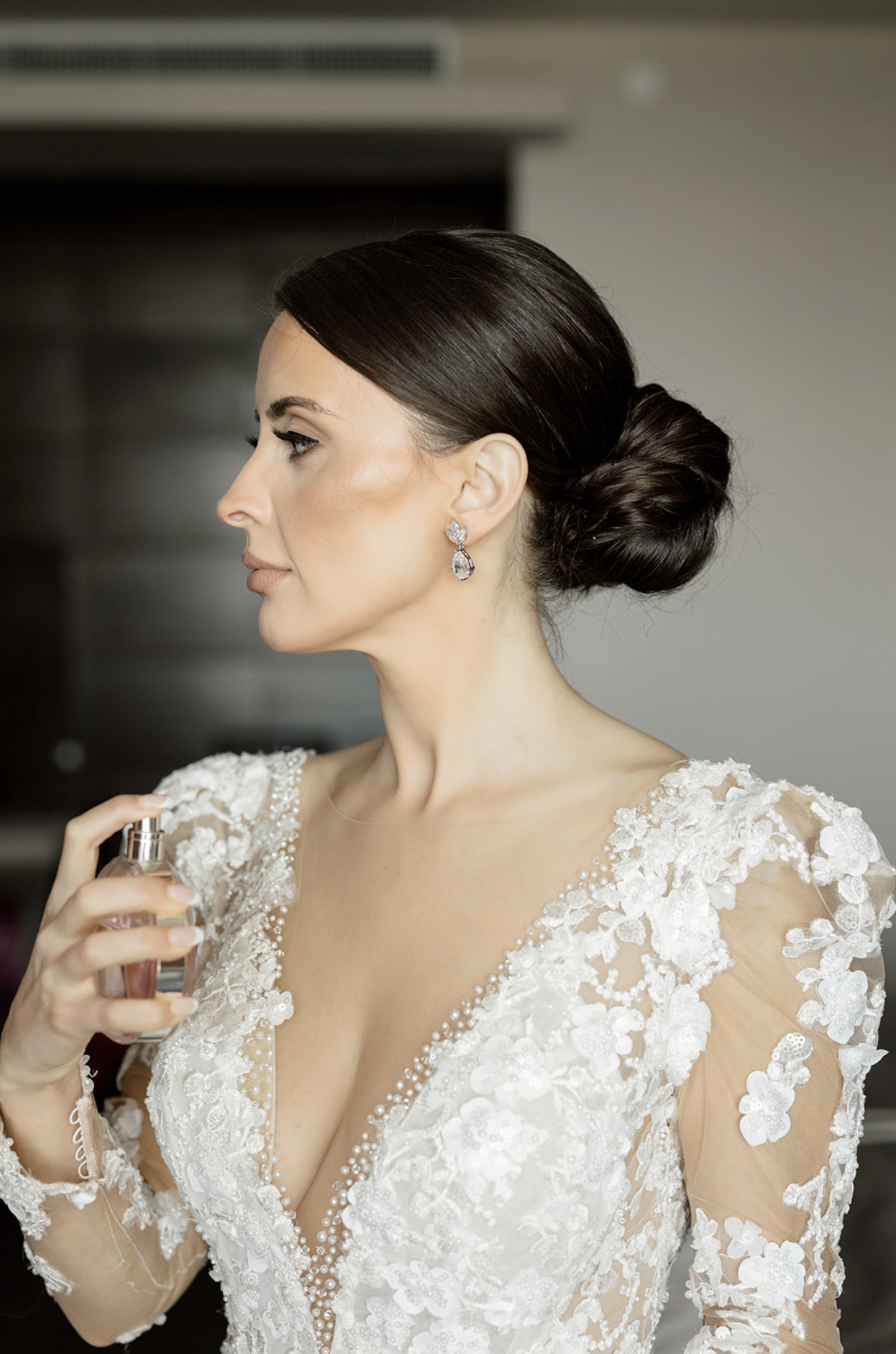 A woman in a white lace wedding dress with floral embroidery, holding a perfume spray, with elegant earrings and her hair styled in an updo.