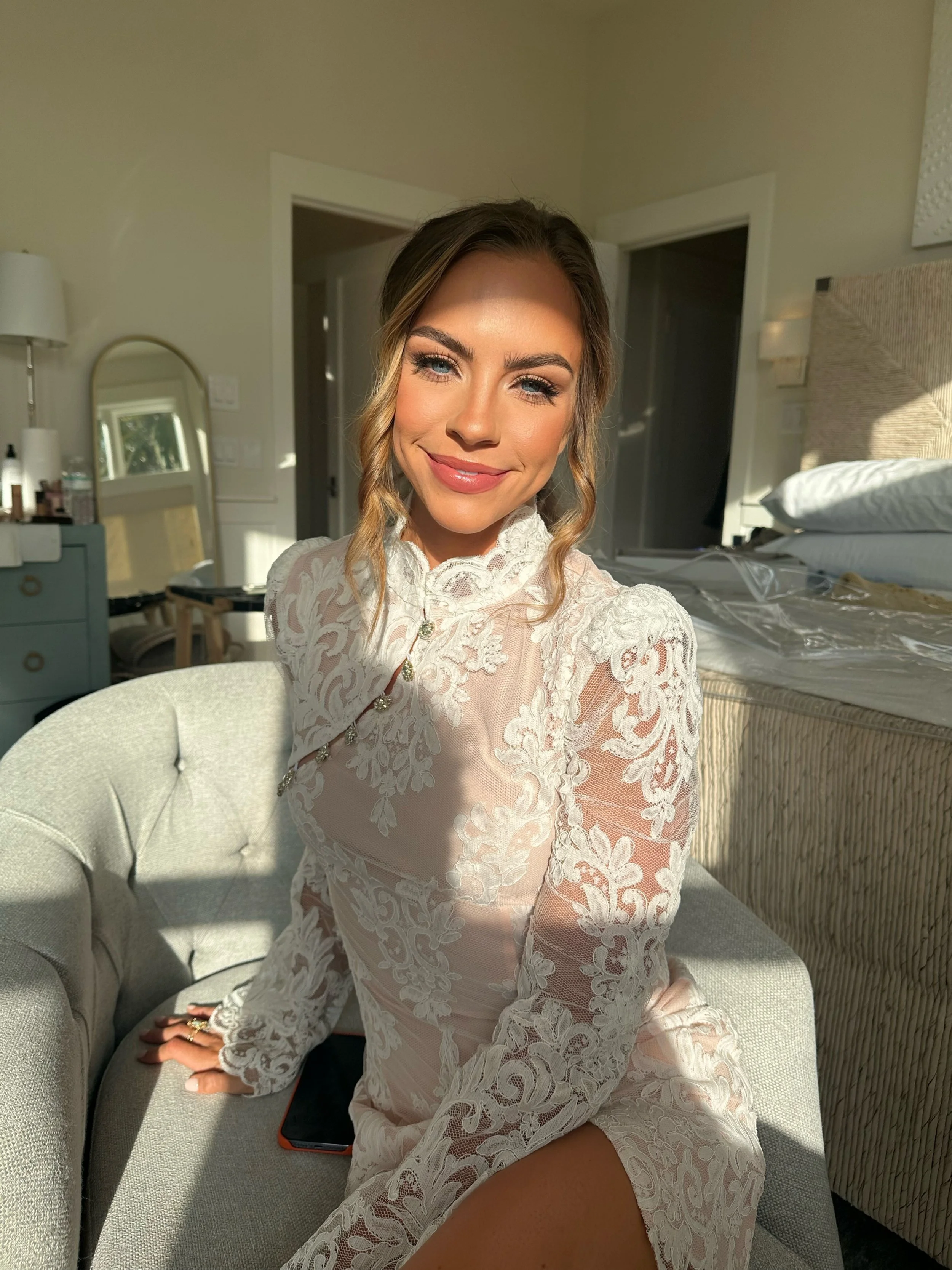 A woman in a lace dress sitting on a light-colored sofa, smiling at the camera with sunlight illuminating her face inside a bedroom.