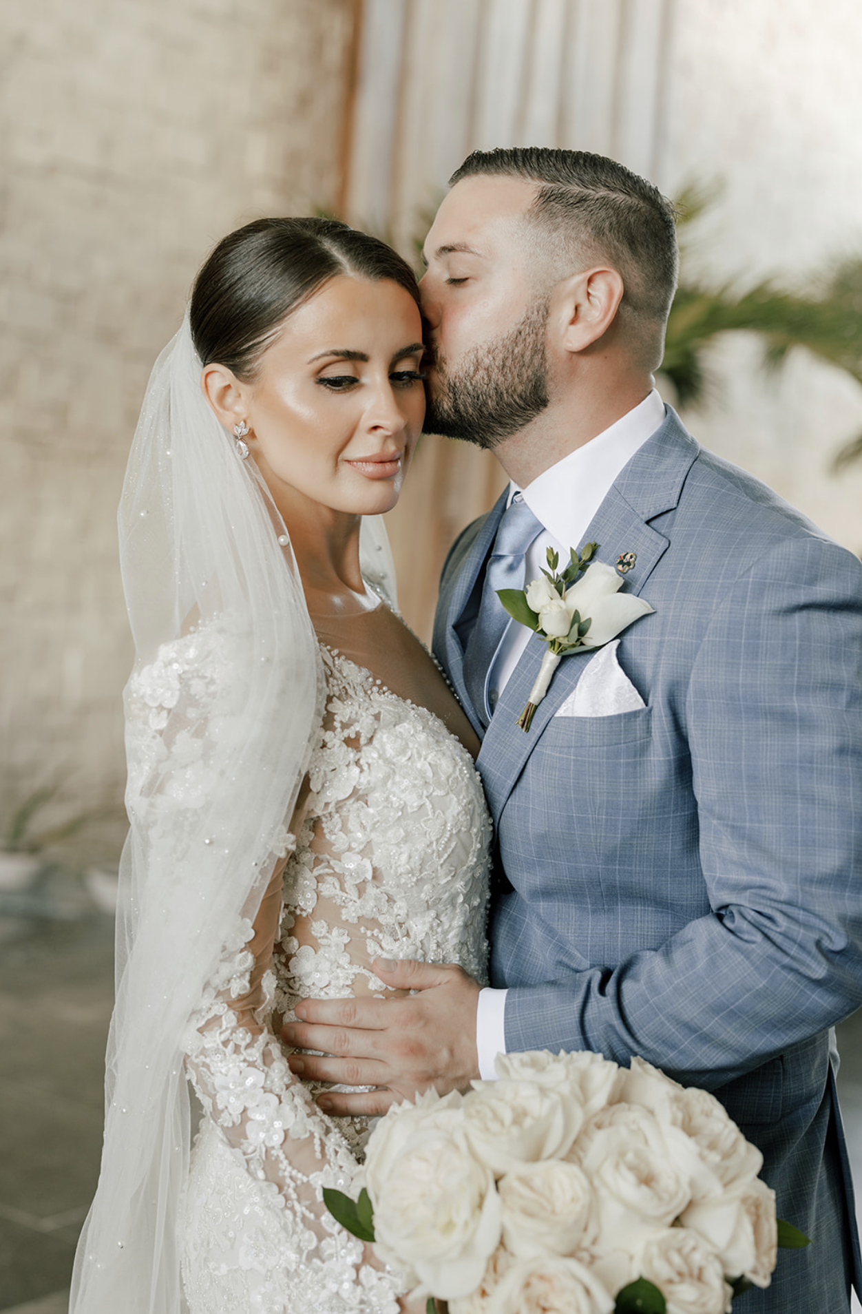 A bride and groom in wedding attire at their wedding, with the groom kissing the bride on her forehead, and the bride holding a bouquet of white roses.