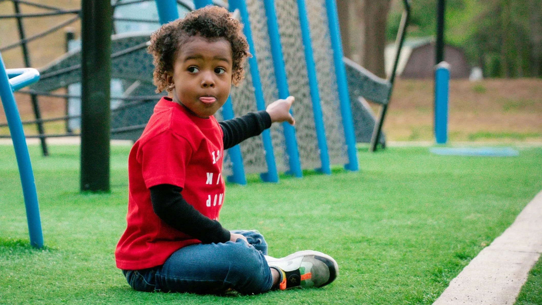 Beautiful curly haired brown skinned little boy sitting on a playground in a red t-shirt while pointing in one direction and looking in another.